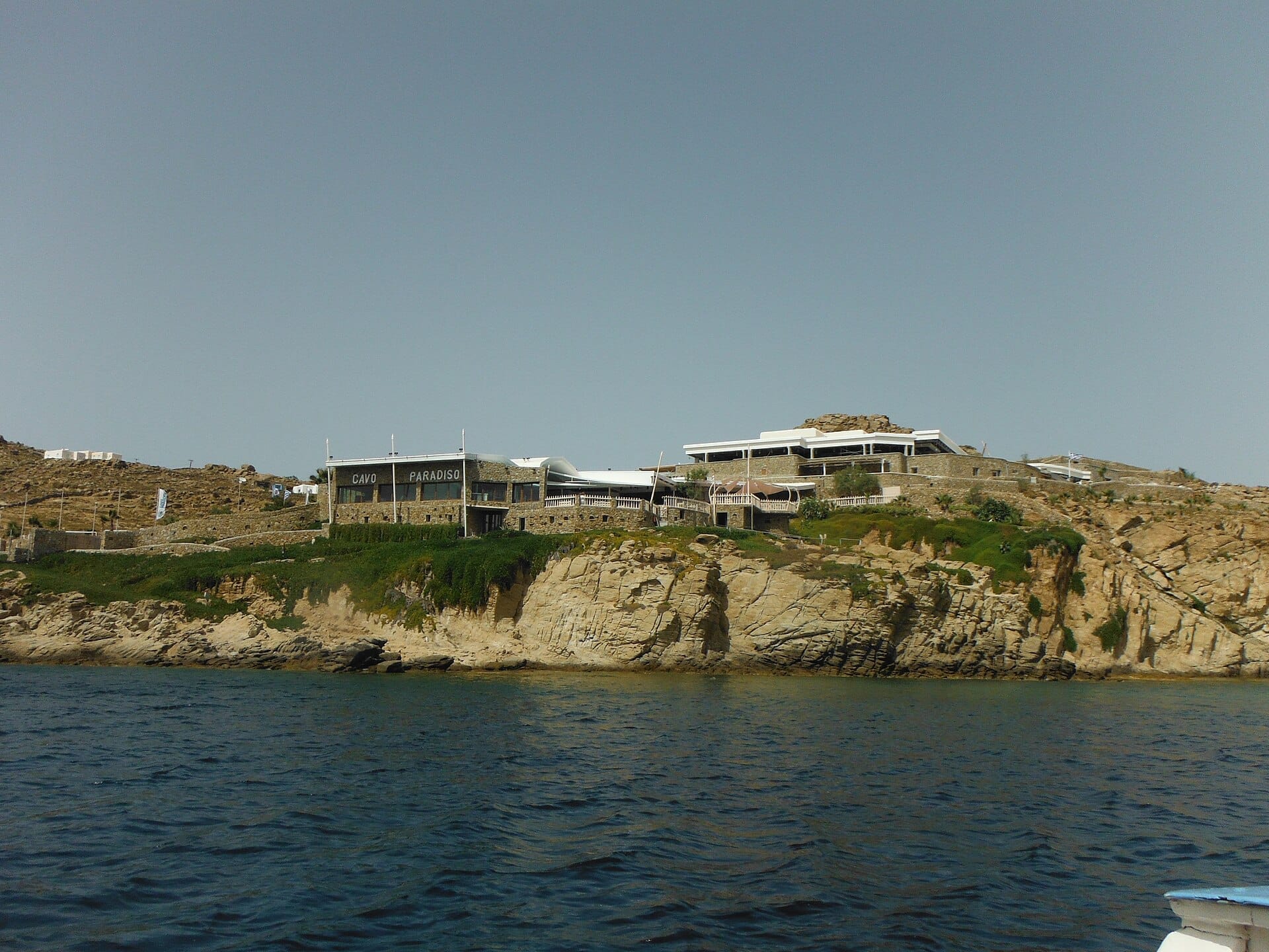Cavo Paradiso Club Mykonos situado en lo alto de un acantilado rocoso sobre el Mar Egeo con sus edificios de piedra y el cielo azul despejado.