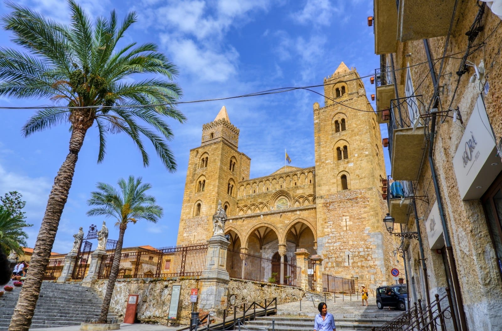 Die Fassade des Doms von Cefalù in Sizilien mit seinen zwei normannischen Türmen, einer Palme und einem dramatisch blauen Himmel, aufgenommen von der Eingangsfreitreppe.