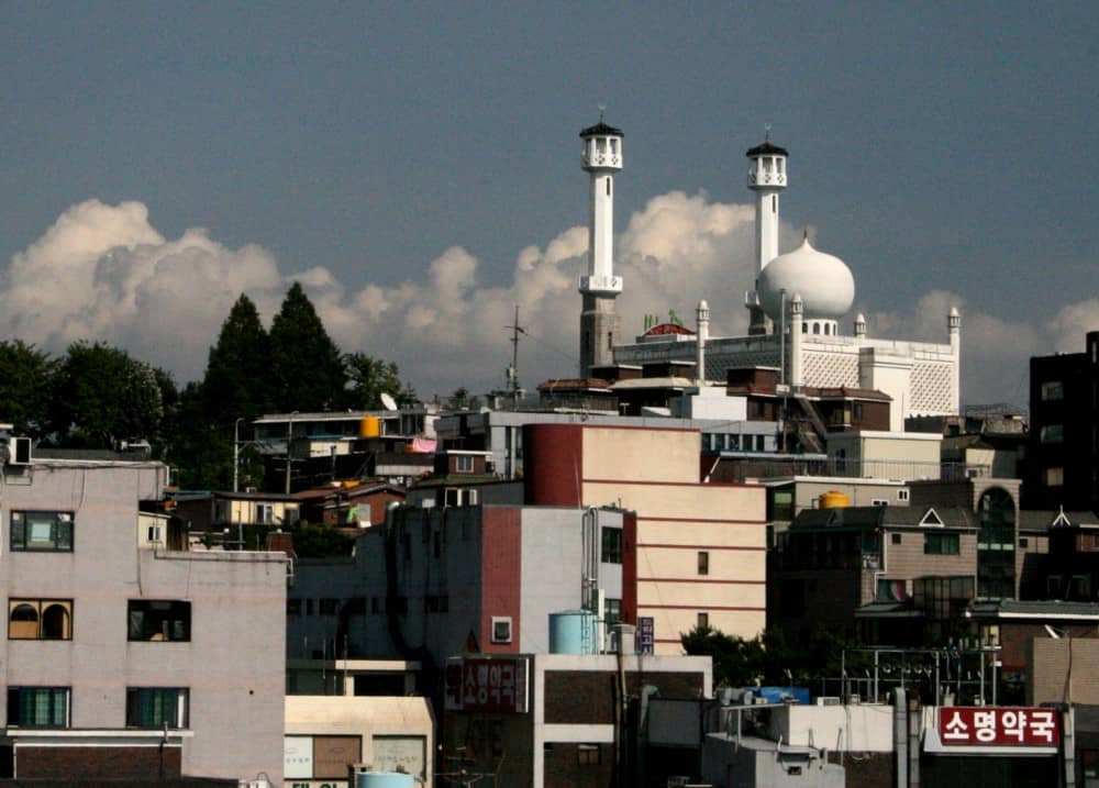 A Mesquita Central de Seul com suas cúpulas brancas e dois minaretes, com vista para os telhados de Itaewon sob um céu parcialmente nublado.