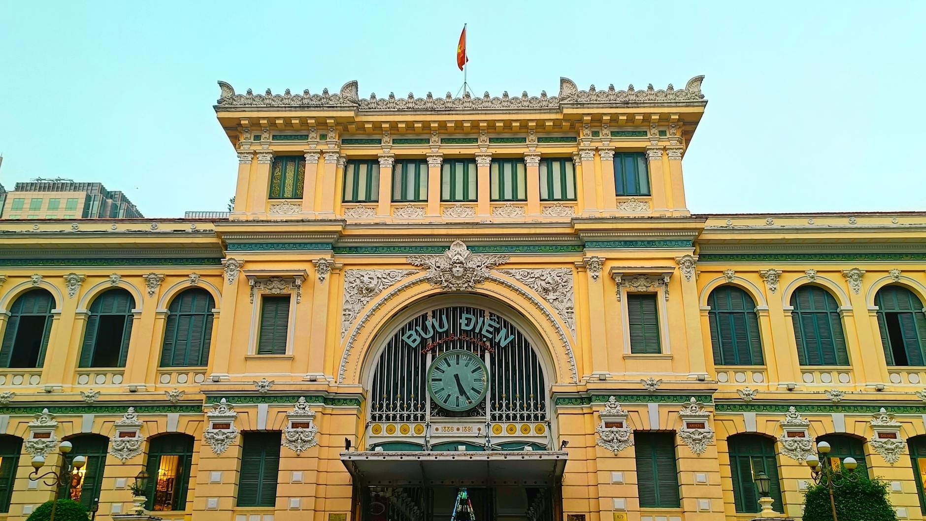 Vista frontal del Correo Central de Saigón, un ornamentado edificio colonial francés de color amarillo con un reloj central y una bandera, bajo un cielo despejado.