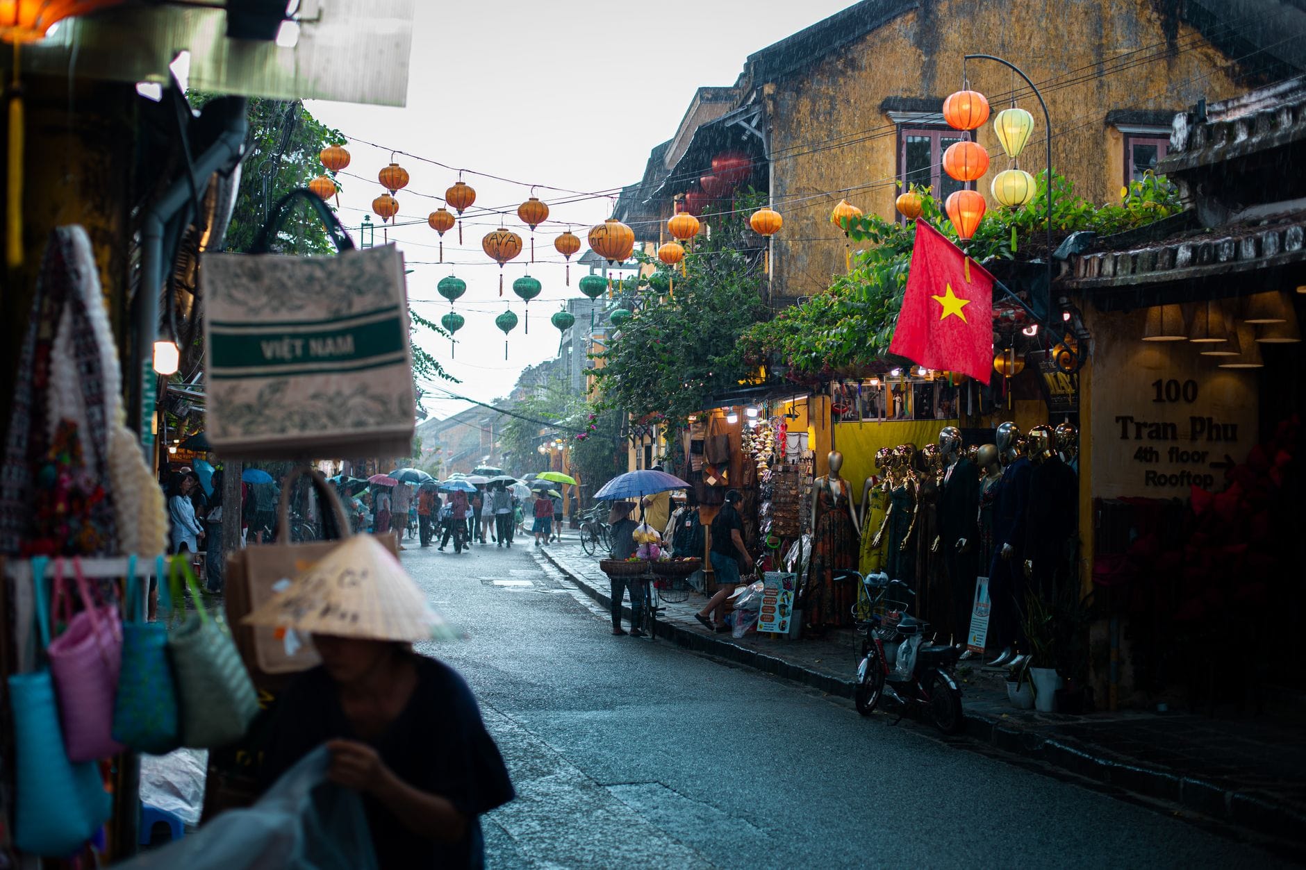 ภาพถนนในวันฝนตกที่เมืองเก่าฮอยอัน พร้อมโคมไฟ ร้านค้า ผู้คนถือร่ม และธงชาติเวียดนามแขวนอยู่ด้านบน