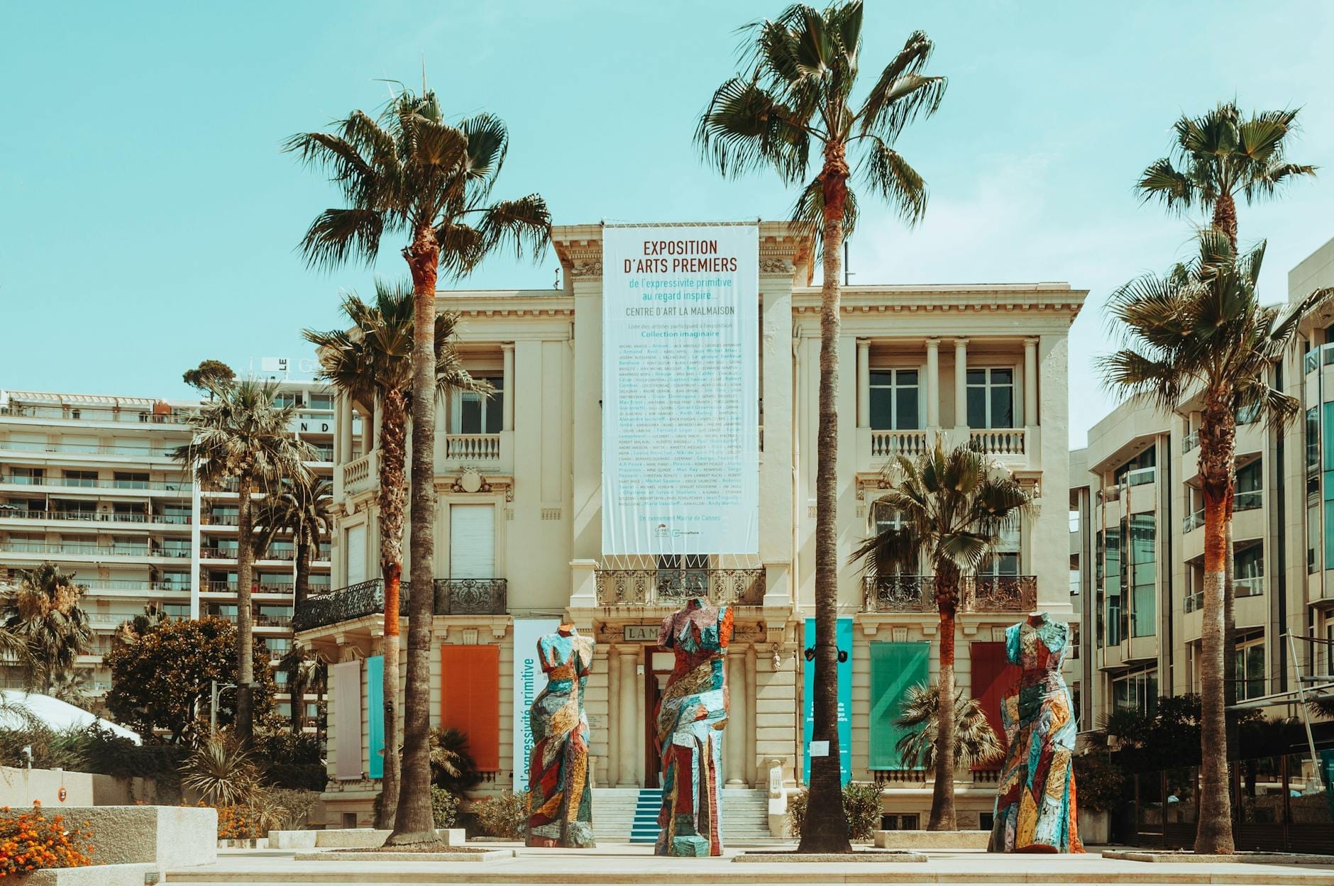 Vista frontale del Centre d'Art La Malmaison a Cannes, fiancheggiato da palme con statue moderne imponenti e striscioni delle mostre, sotto un cielo azzurro luminoso.