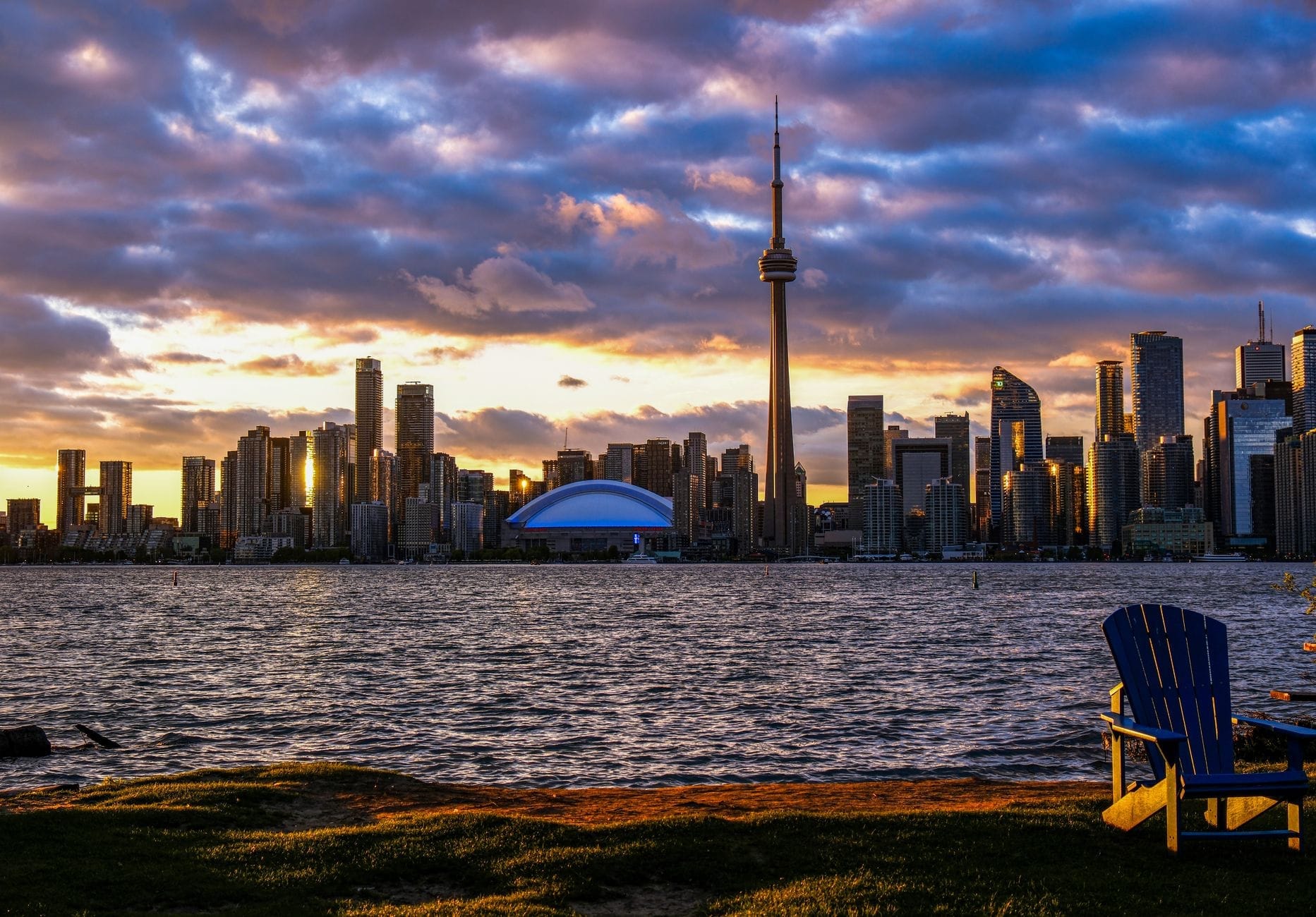 วิวพระอาทิตย์ตกจาก Centre Island เห็นเส้นขอบฟ้าโตรอนโต CN Tower ริมน้ำ และเก้าอี้ Adirondack ว่างอยู่ด้านหน้า