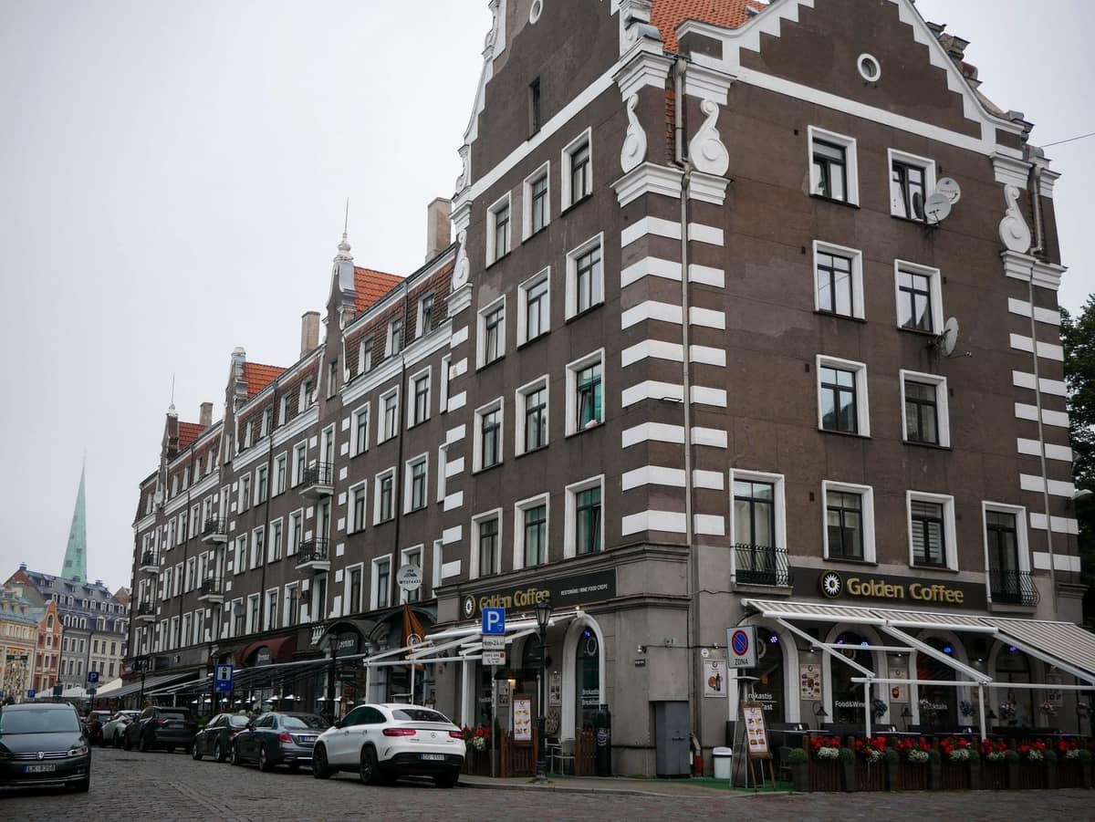 Straßenansicht von Riga Centrs mit historischen Gebäuden, gestreiften Fassaden, Cafés im Erdgeschoss wie Golden Coffee, entlang einer Kopfsteinpflasterstraße geparkten Autos unter bewölktem Himmel.