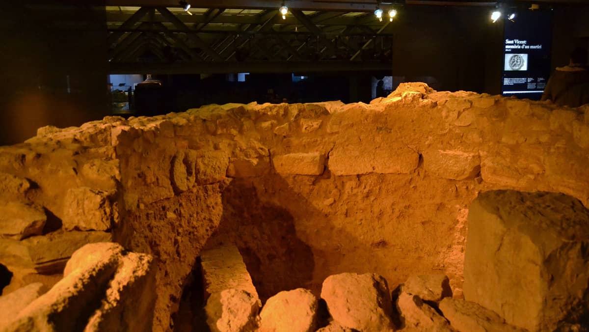 Resti archeologici di antichi muri in pietra e fondamenta all'interno del Centre Arqueològic de l'Almoina, debolmente illuminati con pannelli espositivi sullo sfondo.