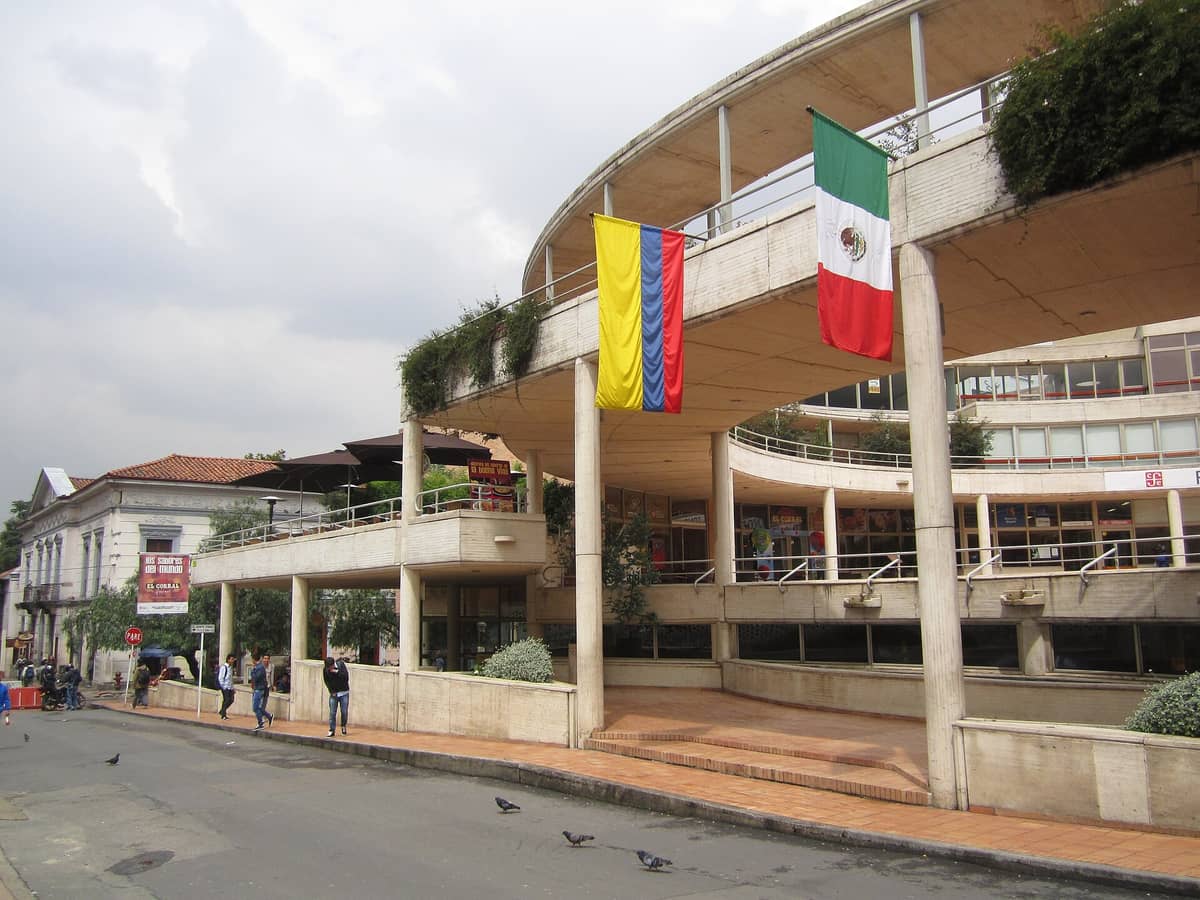 View of the curved exterior of Centro Cultural Gabriel García Márquez, with Colombian and Mexican flags, rounded walkways, and people near the entrance on a cloudy day.