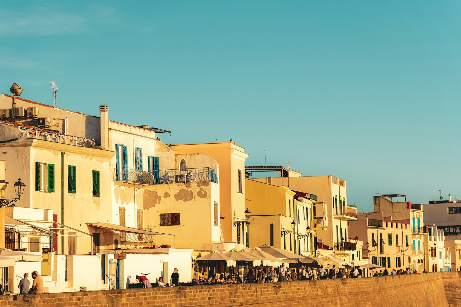 Sonnenbeschienene honigfarbene Gebäude mit blauen Fensterläden säumen die alten Stadtmauern des Centro Storico di Alghero unter einem klaren Himmel, Menschen versammeln sich unten.