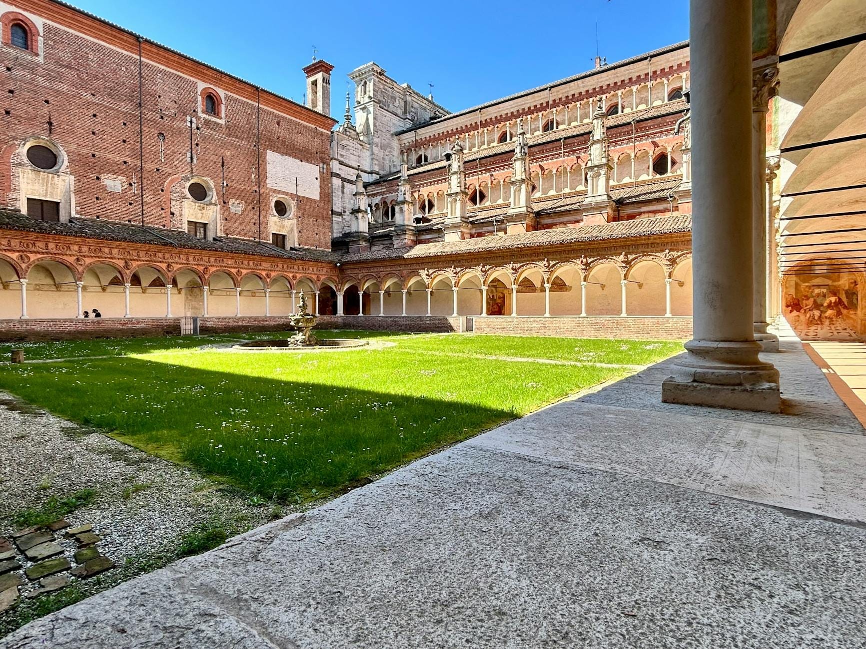 Vista del tranquillo chiostro della Certosa di San Martino con portici ad arco, una fontana centrale e storici muri in mattoni sotto un cielo azzurro limpido.
