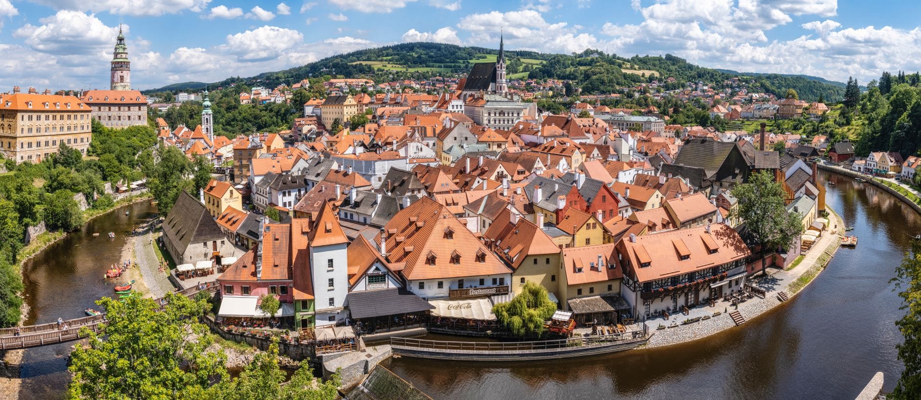 Vista panoramica del centro storico di Český Krumlov con il fiume Moldava e il complesso castellano sotto un cielo soleggiato e luminoso, circondato da una lussureggiante vegetazione.