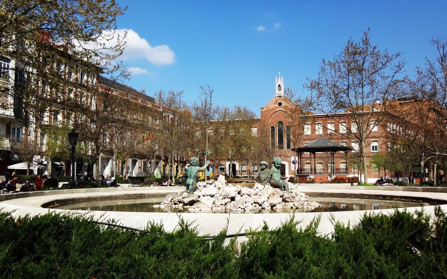 Plaza de Olavide ที่เต็มไปด้วยแสงแดดใน Chamberí กรุงมาดริด โดดเด่นด้วยน้ำพุกลางลานที่มีรูปปั้นสำริด ล้อมรอบด้วยต้นไม้ ม้านั่ง และอาคารอพาร์ตเมนต์สไตล์คลาสสิกในศตวรรษที่ 19