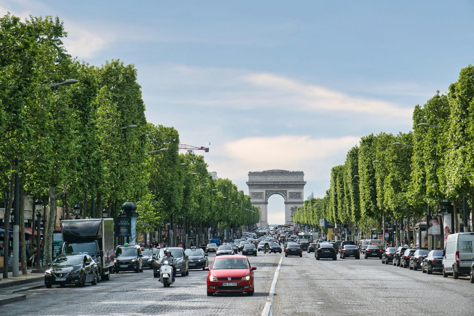 Vista ampia degli Champs-Élysées verso l'Arco di Trionfo, fiancheggiati da alberi verdi e animati dal traffico sotto un cielo luminoso.