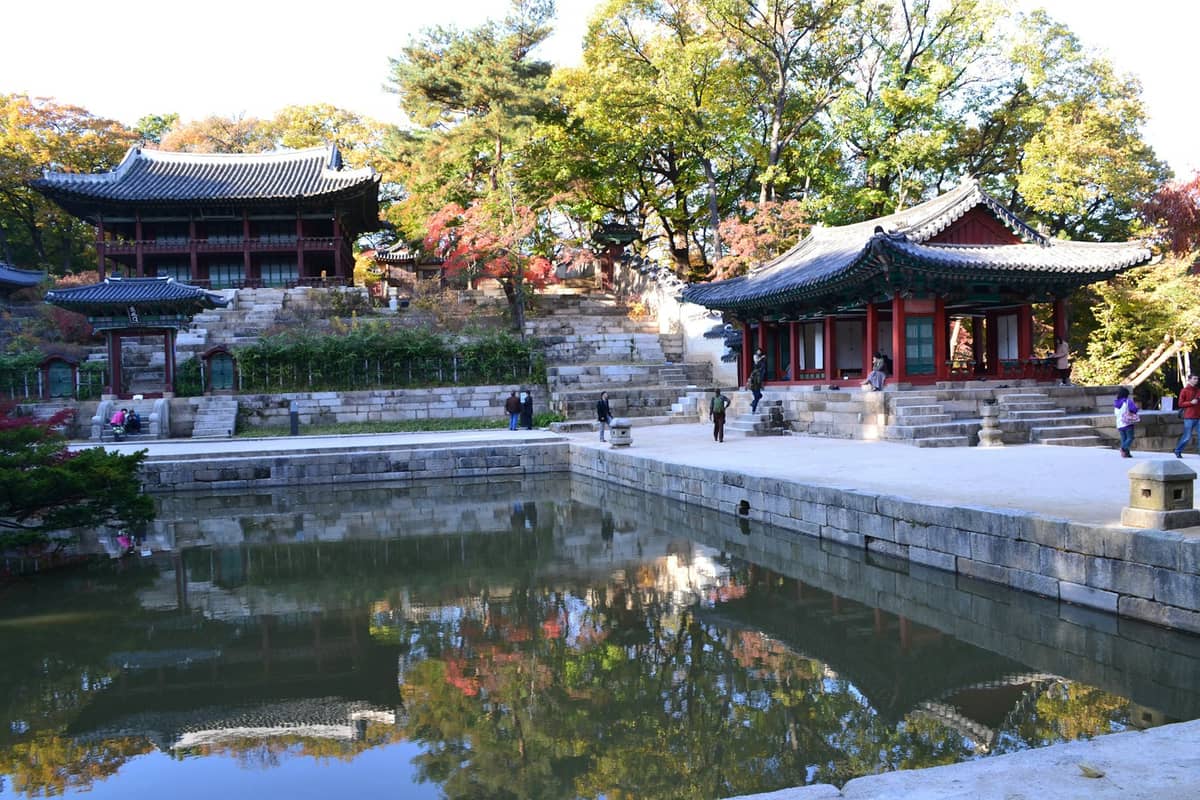 Vista ampla do Palácio Changdeokgung e do lago do Jardim Secreto com pavilhões tradicionais, árvores frondosas e reflexos na água em um dia ensolarado.