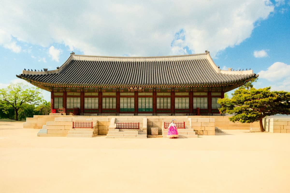 Vista ampla do Salão Myeongjeongjeon no Palácio Changgyeonggung, em Seul, com arquitetura tradicional do estilo Joseon sob um céu azul e uma pessoa vestindo hanbok.