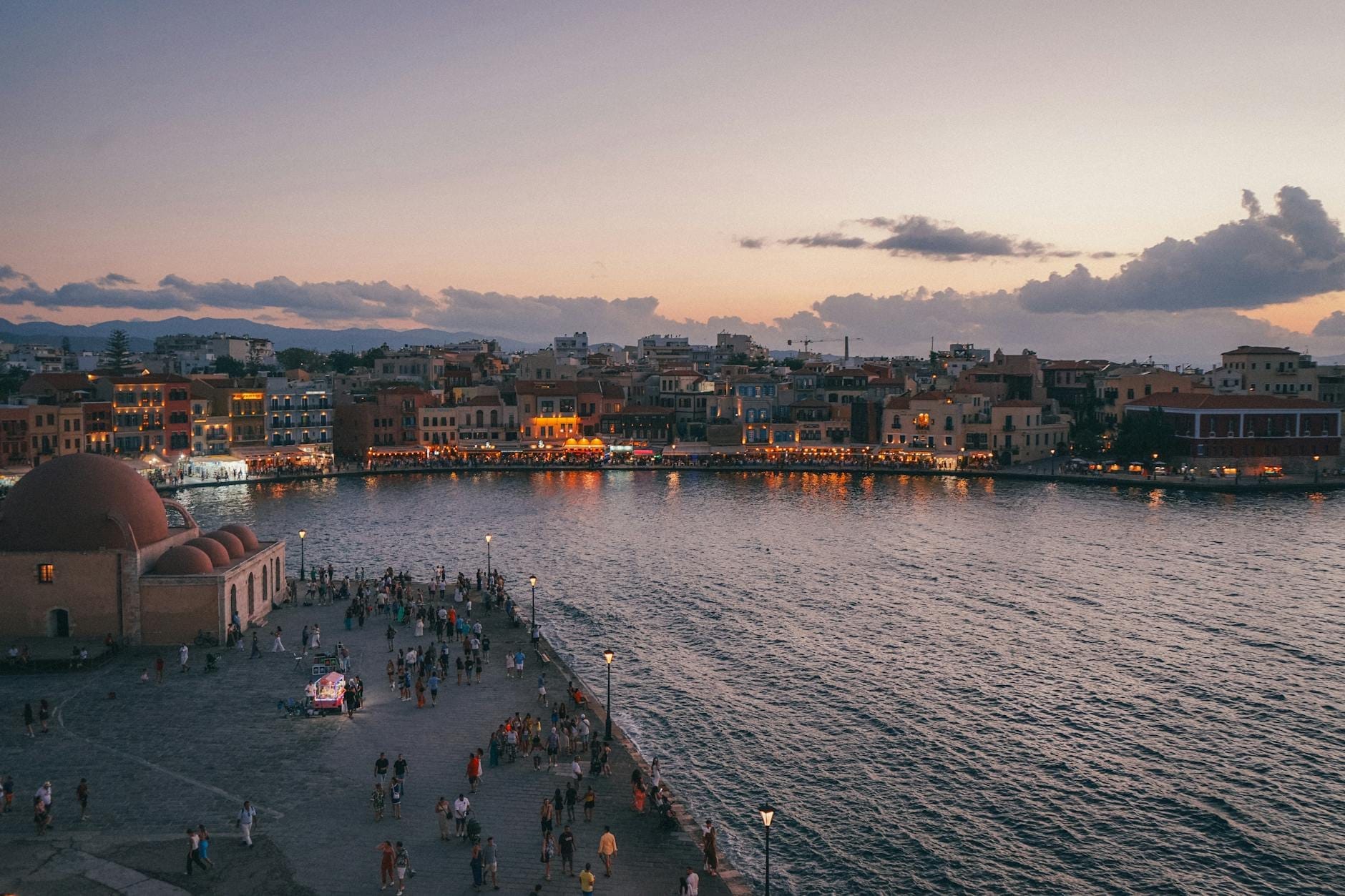 Vista serale del porto della Città Vecchia di Canea con i palazzi veneziani, la moschea a cupola e l'animato lungomare, illuminati sotto un cielo colorato al tramonto.