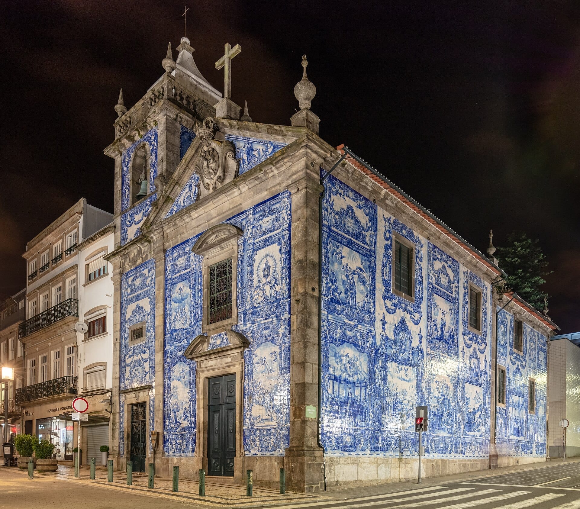 La Capela das Almas a Porto illuminata di notte, con le sue vivaci piastrelle azulejo blu e bianche e i dettagli architettonici barocchi su un angolo di strada.