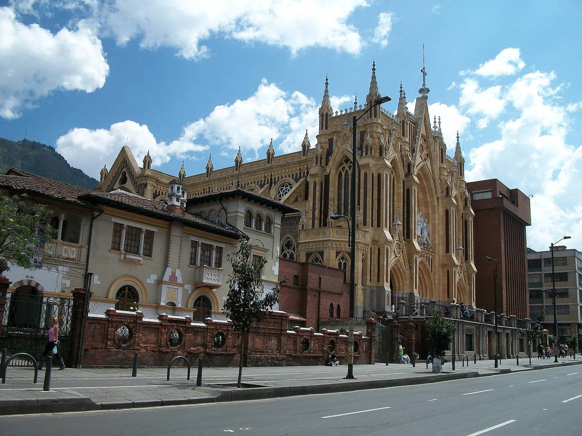 Ornate Gothic-style cathedral and historic buildings on a quiet street in Chapinero, Bogotá, under a bright blue sky with scattered clouds.