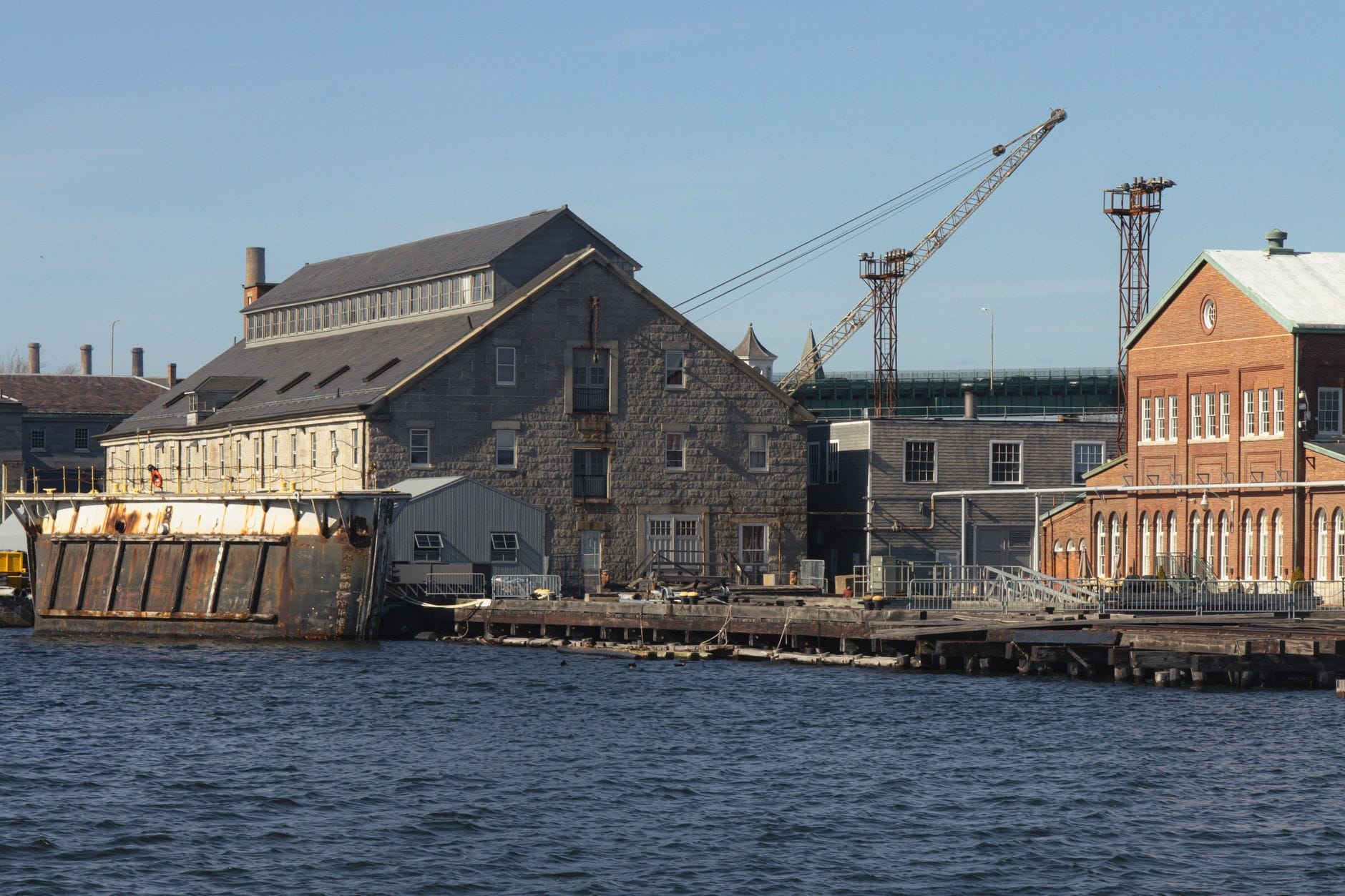 Edifici storici in pietra e mattoni sul lungomare del Charlestown Navy Yard, con una gru e una chiatta ormeggiata, simbolo del suo passato marittimo e industriale.