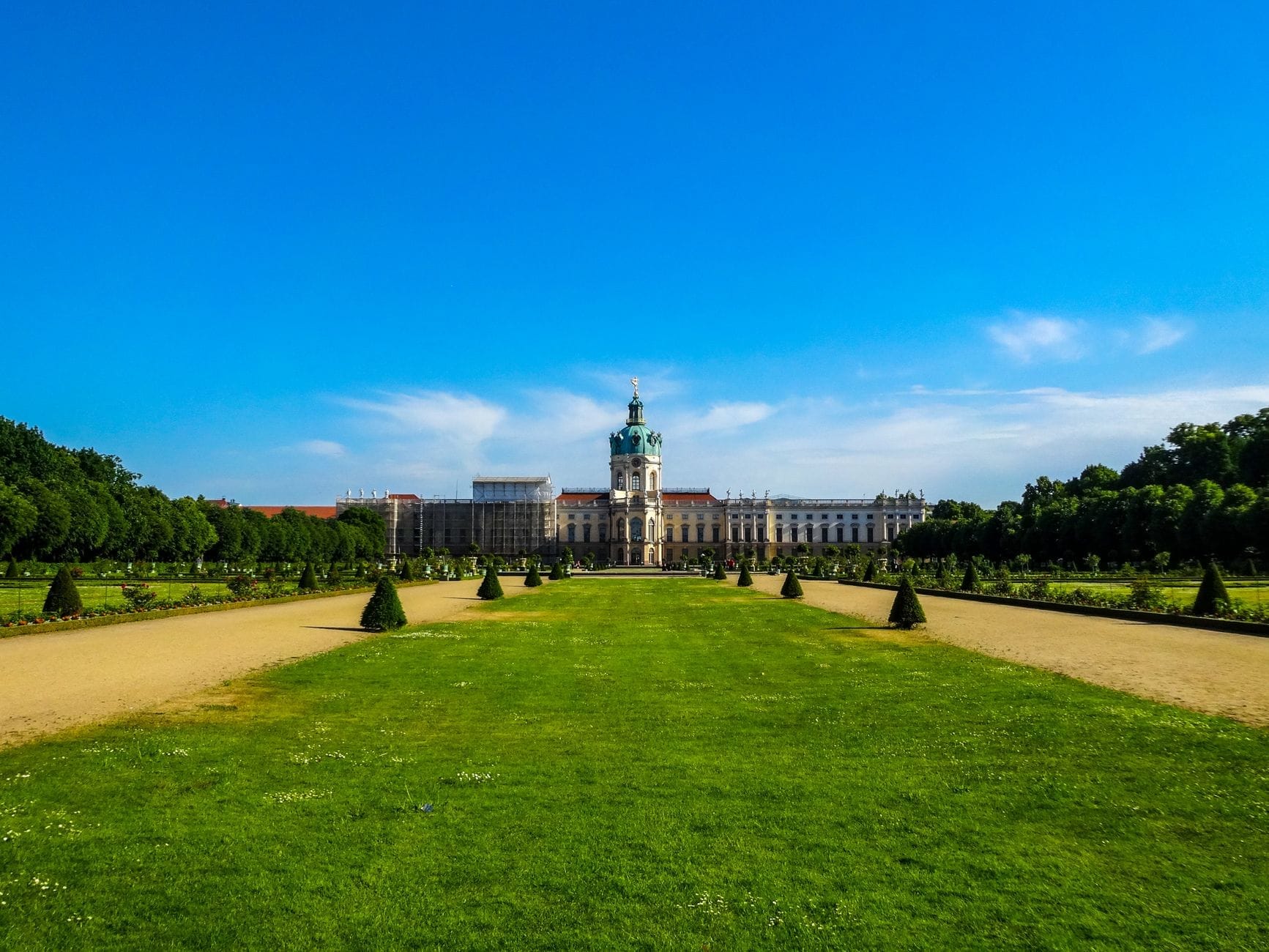 Vista panoramica del Palazzo di Charlottenburg con i giardini formali verdi, i vialetti simmetrici e un cielo azzurro brillante, con l'intera facciata del palazzo in fondo al giardino.