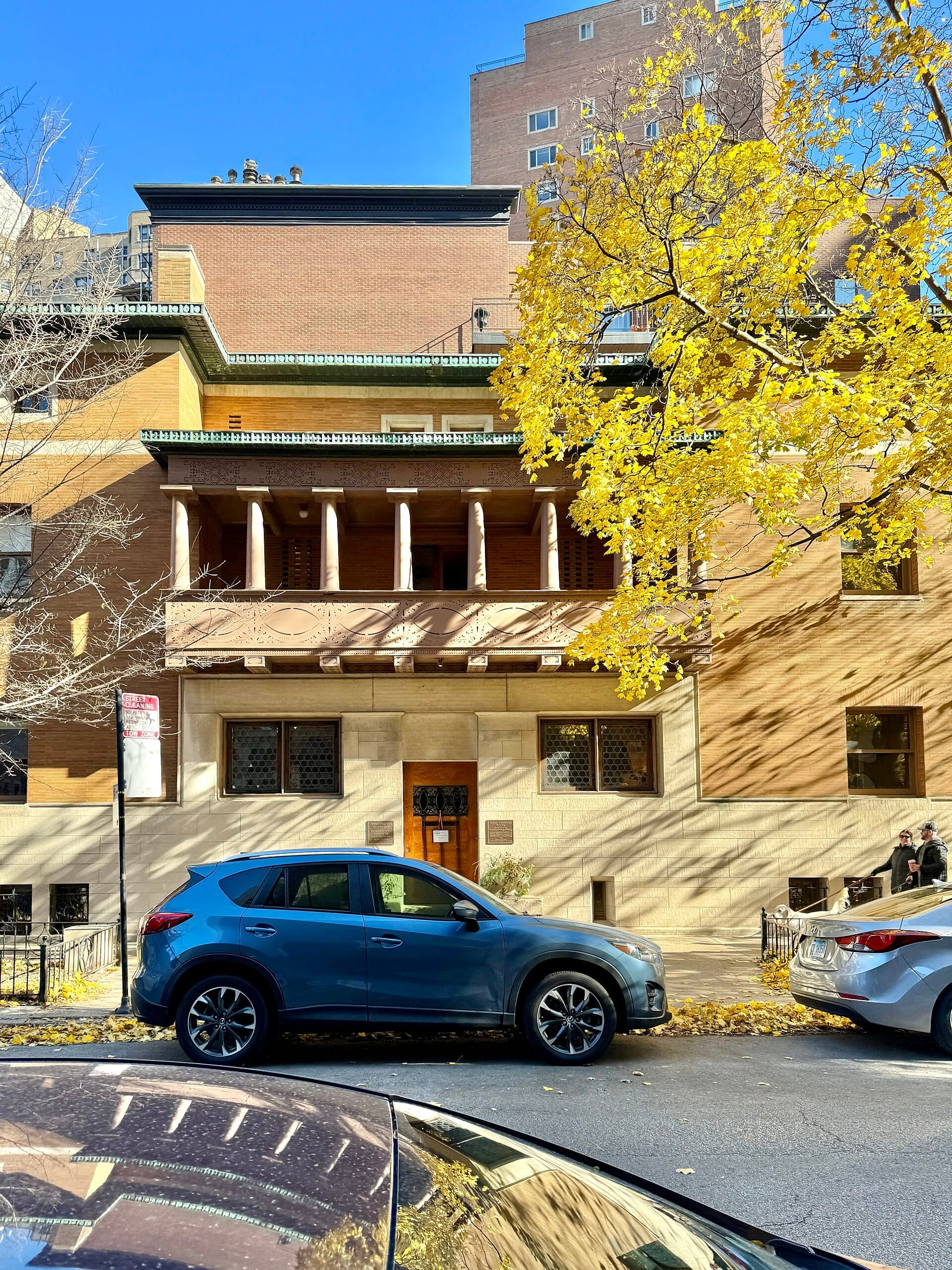 Vista frontale del Charnley-Persky House Museum con alberi autunnali, classica facciata in mattoni e pietra, e auto parcheggiate lungo la strada.