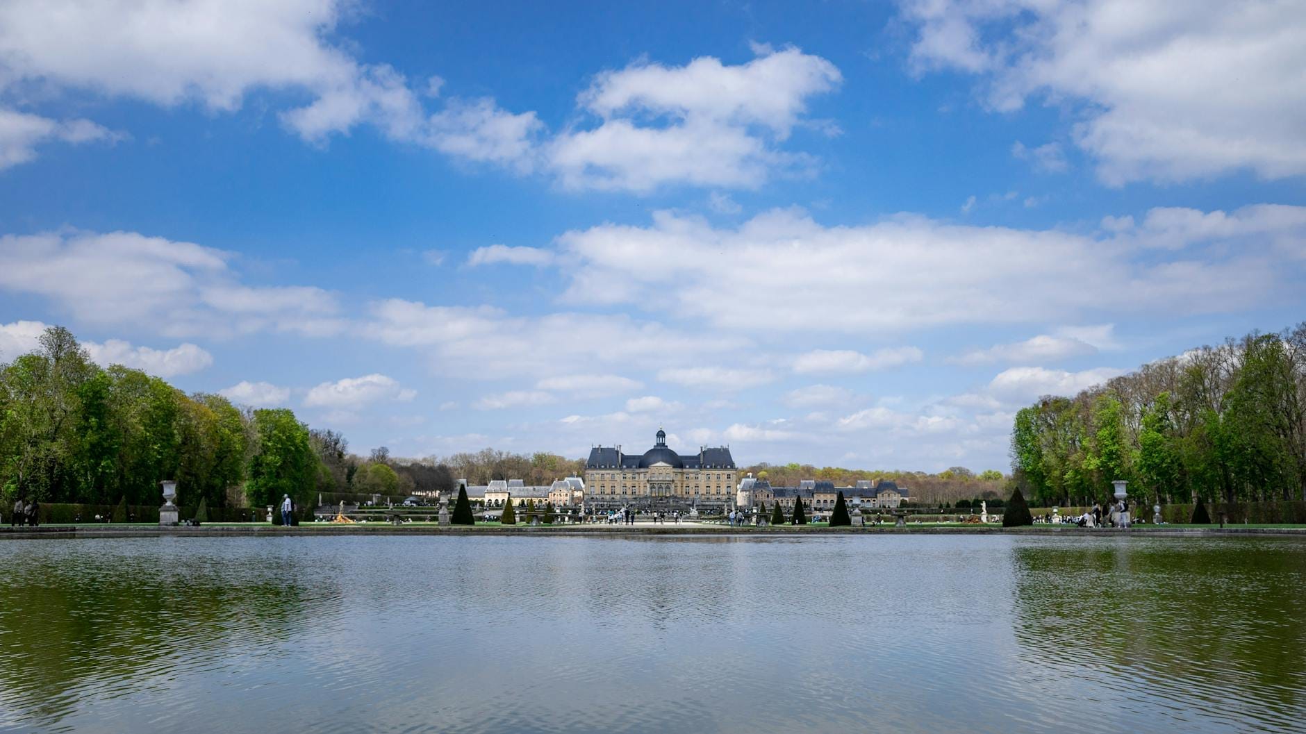 Il Château de Vaux-le-Vicomte visto dall'altro lato di un ampio specchio d'acqua, circondato da giardini formali sotto un cielo azzurro con qualche nuvola sparsa.