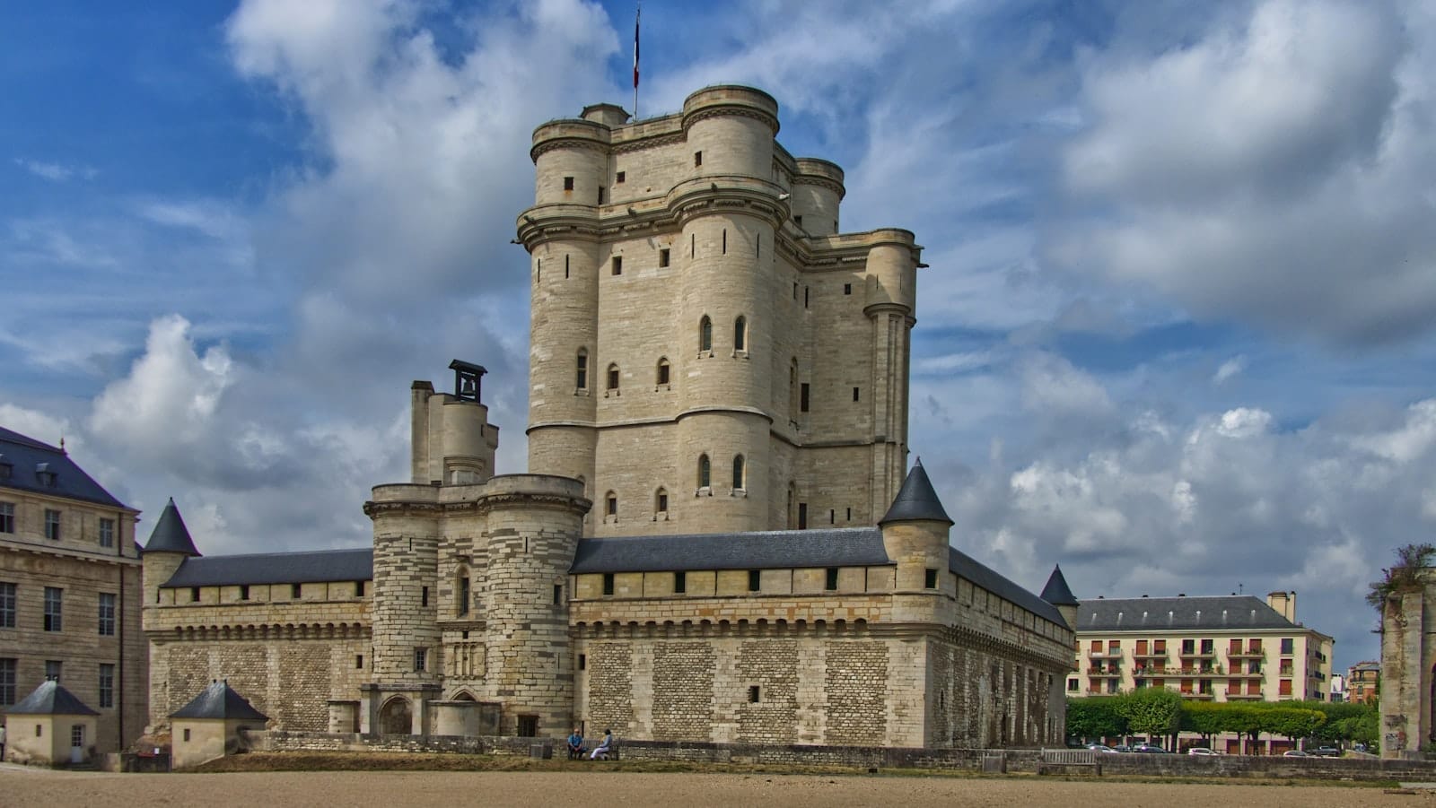 Vista panoramica del Château de Vincennes con il suo imponente mastio medievale, le torri merlate e le mura della fortezza sotto un cielo blu intenso con qualche nuvola sparsa.
