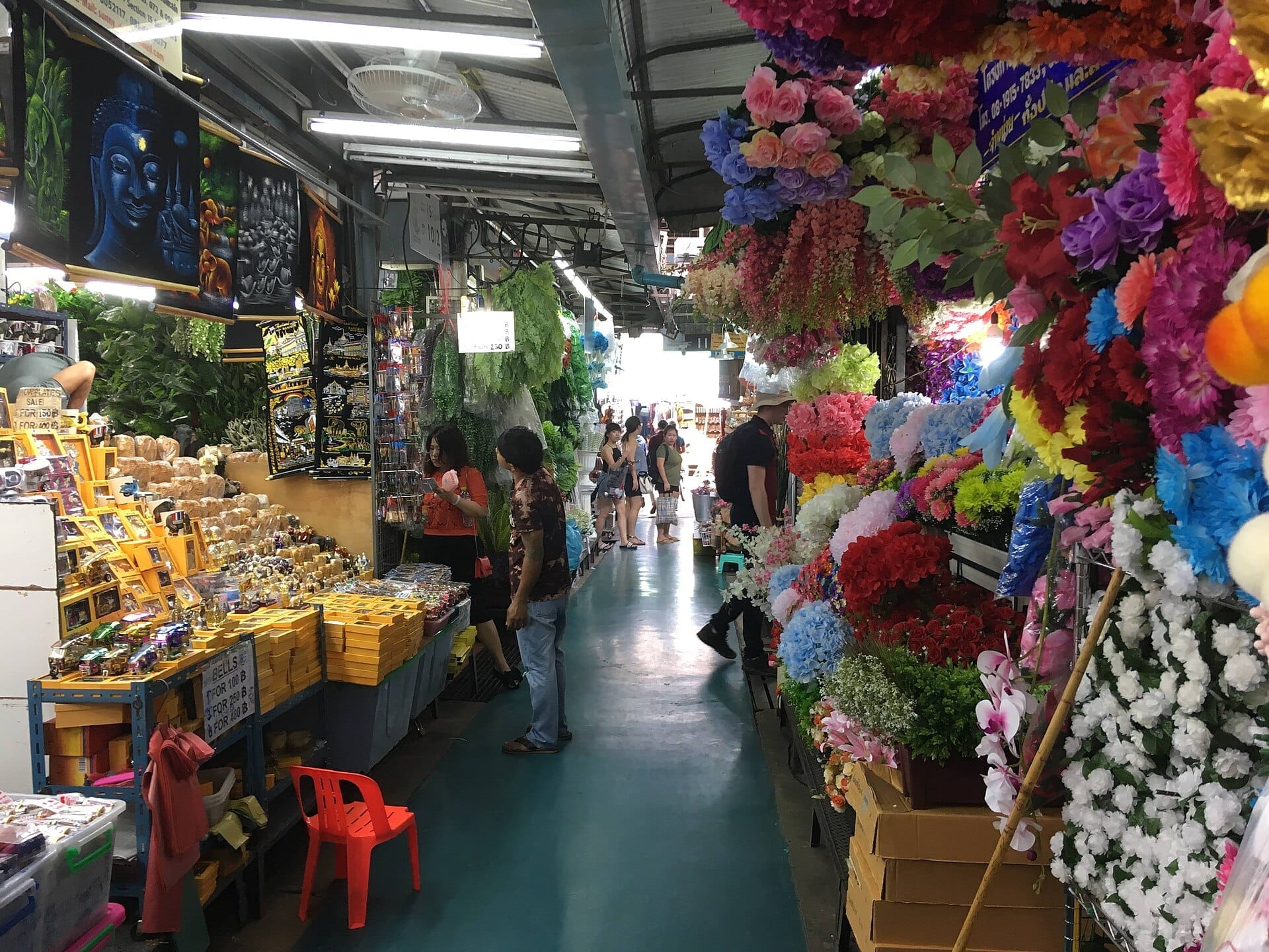 Corridoio interno del Chatuchak Weekend Market con fiori artificiali colorati, bancarelle di souvenir e visitatori che curiosano a Bangkok
