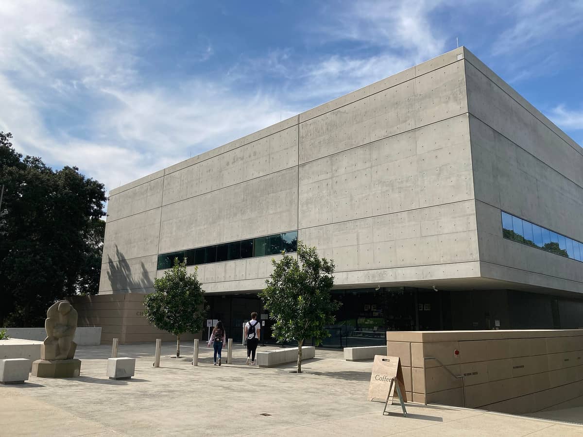 Vista exterior del Chau Chak Wing Museum en la Universidad de Sídney, con el moderno edificio de hormigón, la entrada y personas en los alrededores.