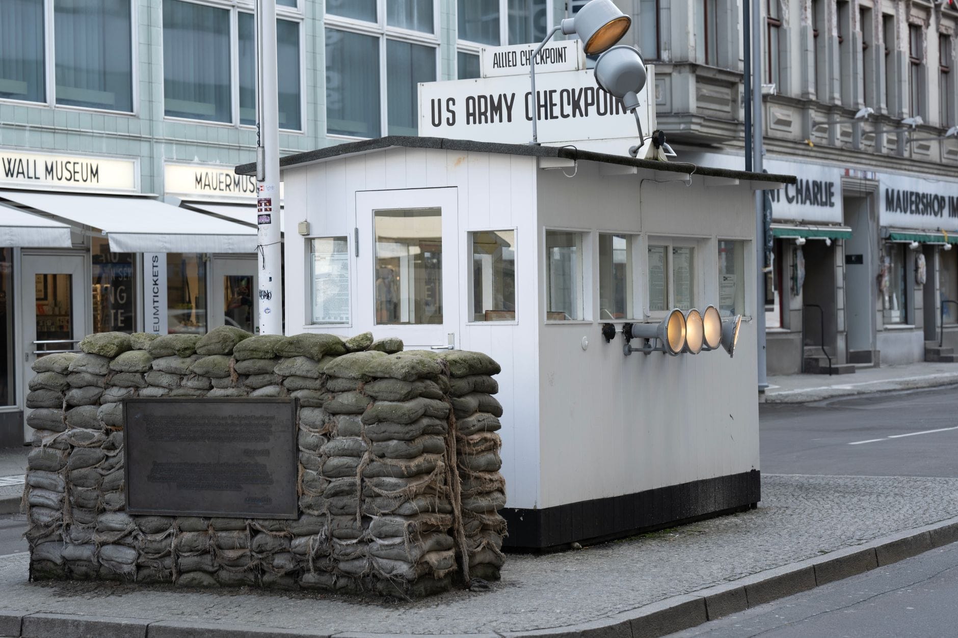 La guardiola ricostruita di Checkpoint Charlie con il cartello dell'esercito americano e i sacchi di sabbia, su una strada di Berlino con il museo e i negozi sullo sfondo.