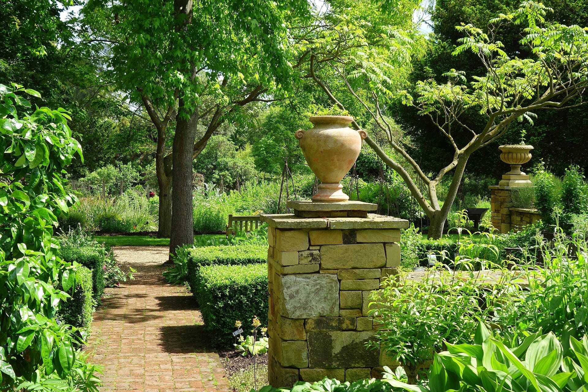 Vista soleggiata del Chelsea Physic Garden con sentieri in pietra, alberi frondosi, rigogliosa vegetazione e urne classiche su piedistalli in pietra.