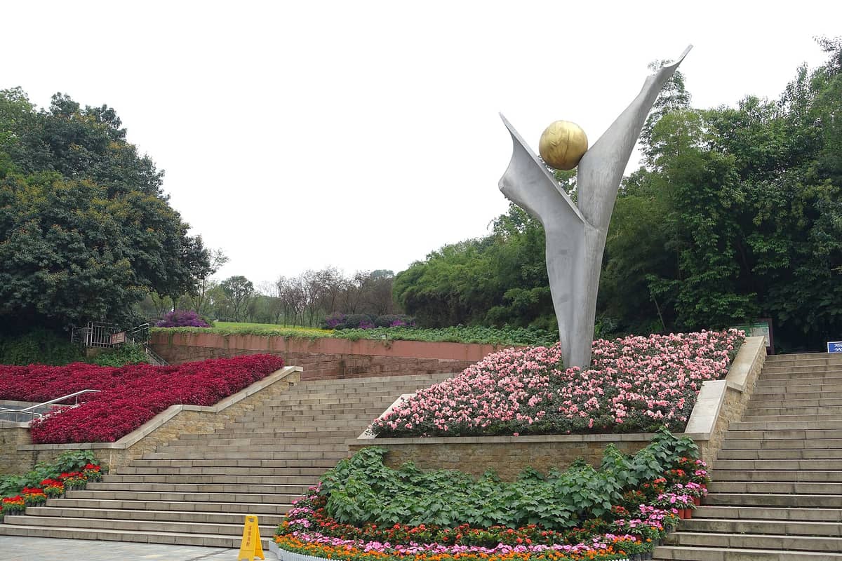 Weitläufiger Blick auf den Haupteingang des Botanischen Gartens Chengdu mit einer markanten silber-goldenen abstrakten Skulptur, umgeben von Blumenbeeten und üppigem Grün.