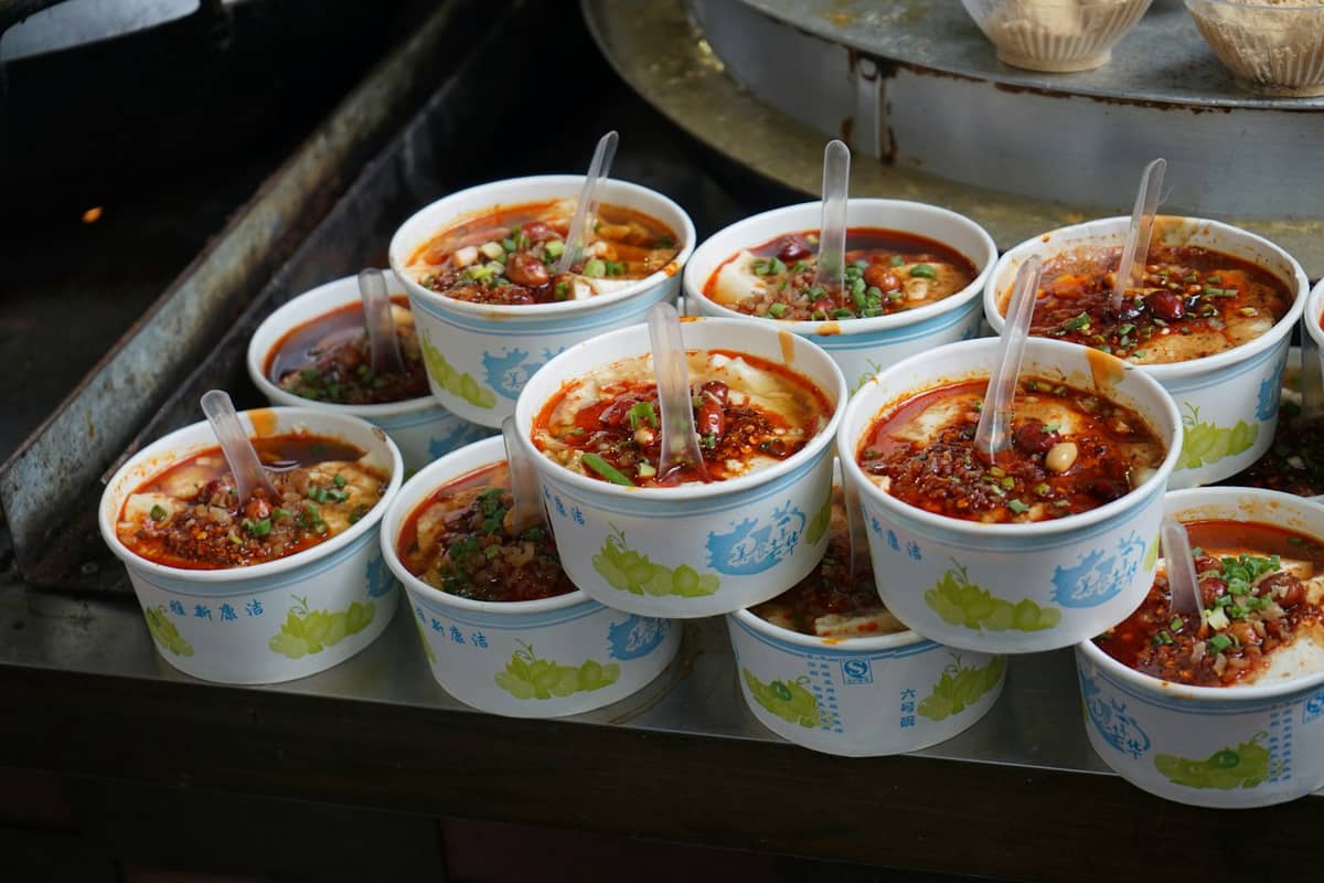 Filas de comida callejera picante de Sichuan servida en cuencos con aceite de chile, cacahuetes y hierbas en un puesto de mercado de Chengdu.