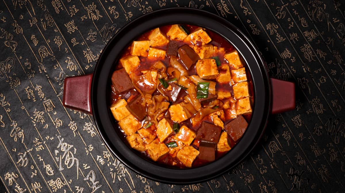 Draufsicht auf eine Schüssel Mapo Tofu, ein klassisches scharfes und taubendes Gericht aus Sichuan, auf einem dekorativen Tuch mit chinesischer Kalligrafie.