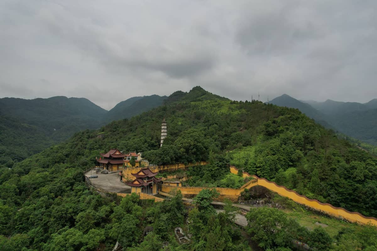 Vista aérea de un complejo de templos taoístas entre colinas verdes exuberantes con un sendero sinuoso y un dramático cielo nublado