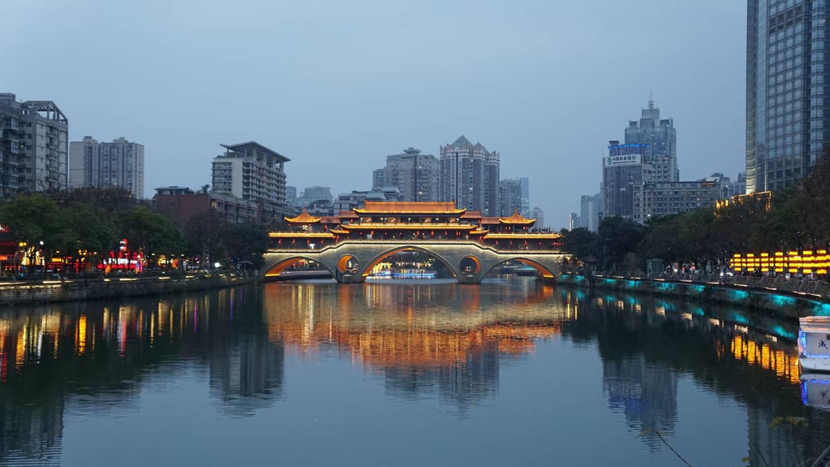 Vista nocturna del icónico Puente Anshun de Chengdu iluminado y reflejado en el río Jin, rodeado de edificios modernos.