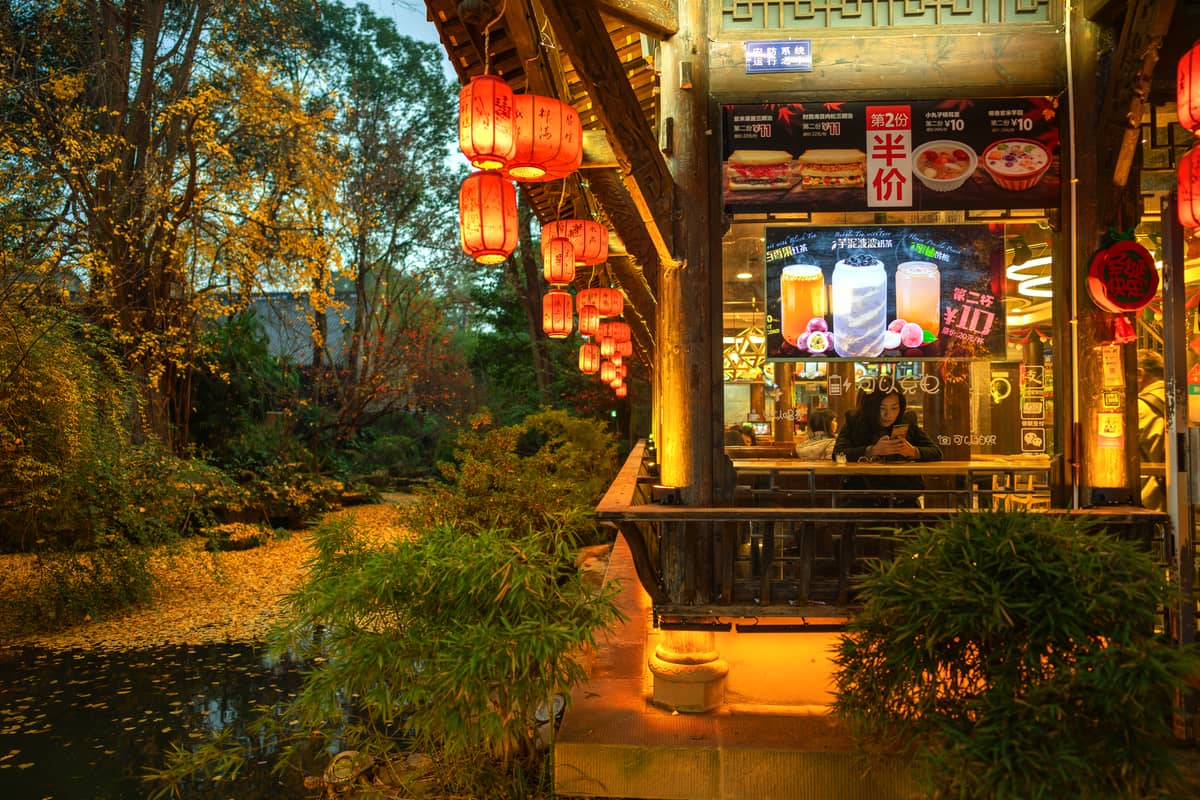 Casa de té china tradicional iluminada con faroles rojos junto a un pequeño lago y exuberante vegetación al atardecer, evocando una atmósfera tranquila y romántica.