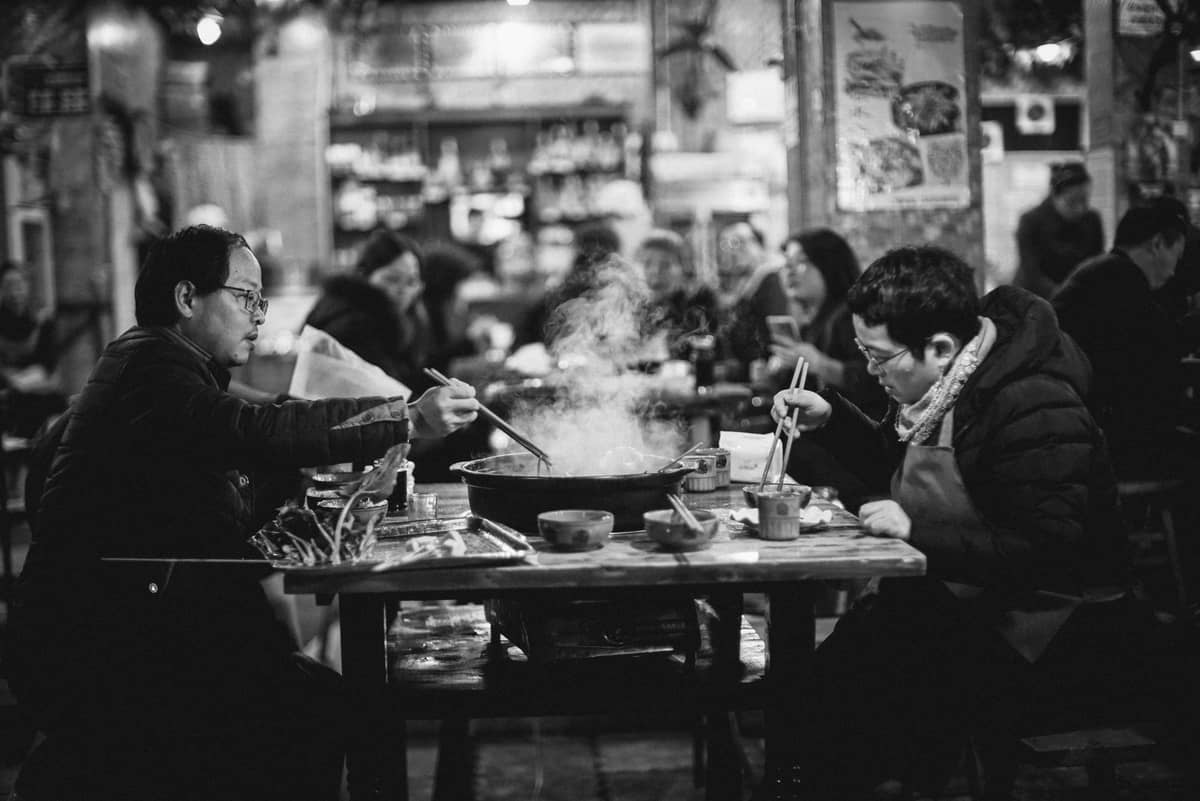Dos personas compartiendo una olla caliente en un animado restaurante, con vapor saliendo de la olla y otros comensales comiendo al fondo.