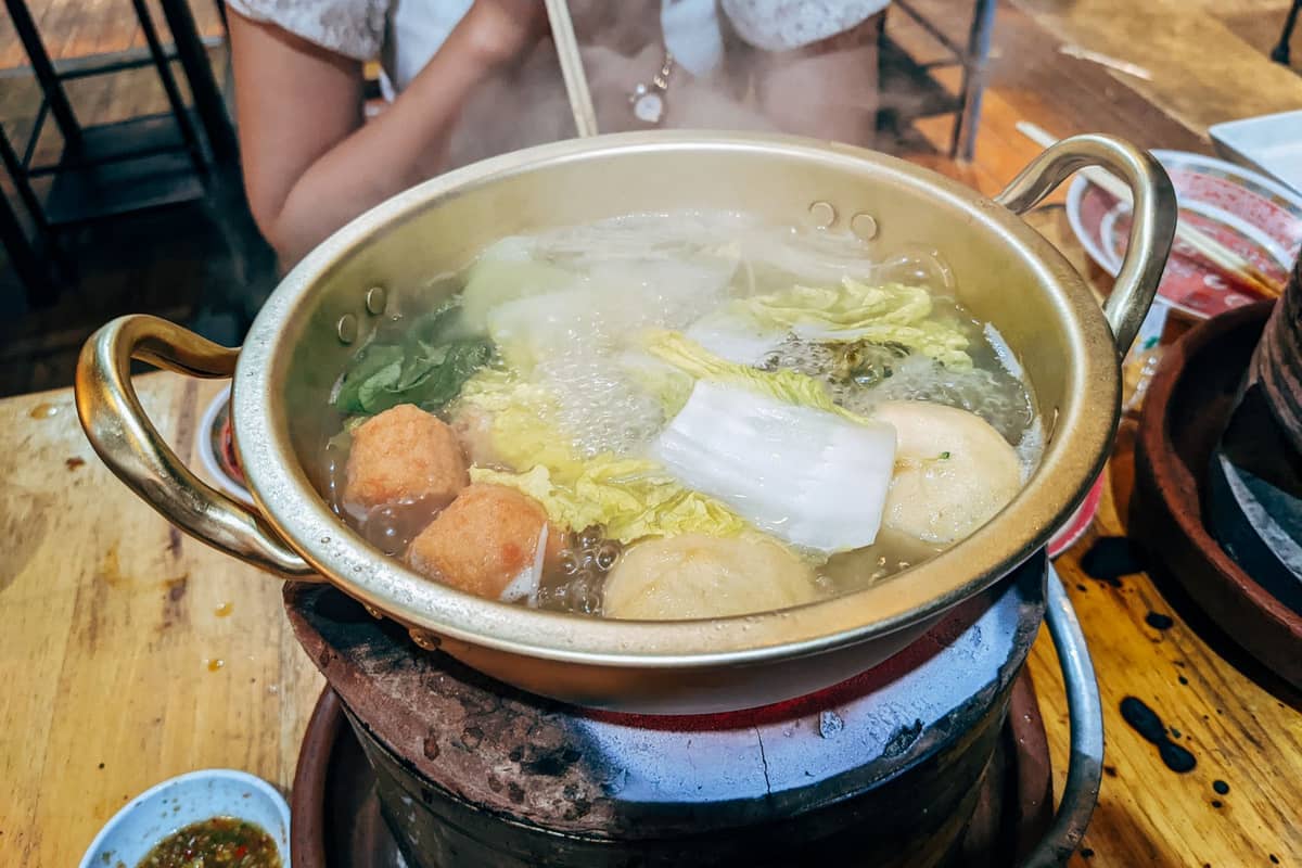 Una pentola fumante di hot pot con verdure e polpette in ebollizione su un fornellino da tavolo, con una persona che usa le bacchette.