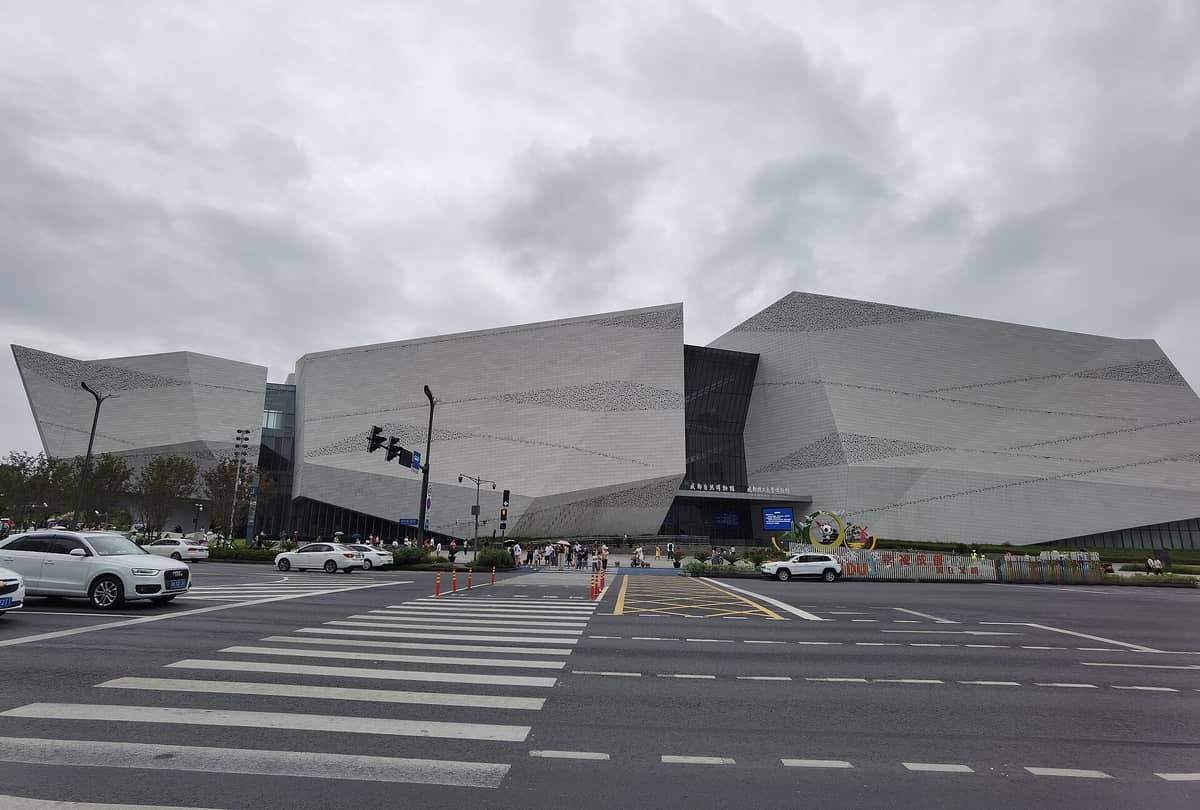 Chengdu Doğa Tarihi Müzesi'nin kendine özgü geometrik mimarisinin sokağın karşısından geniş açıyla görünümü; önde yayalar ve arabalar.