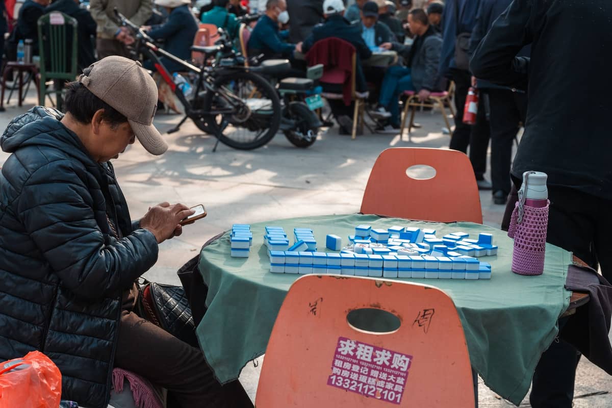 Persone sedute all'aperto ai tavoli a giocare a mahjong in una trafficata area di parco pubblico a Chengdu.