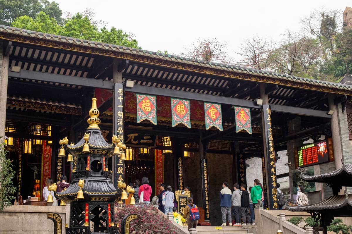Menschen besuchen einen chinesischen buddhistischen Tempel mit bunten Bannern und einem Räuchergefäß am Eingang, umgeben von Grünflächen.