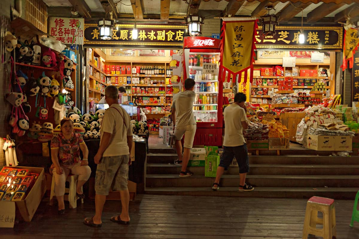 Persone che fanno acquisti in un negozio di souvenir di Chengdu che vende giocattoli panda, snack e prodotti locali sotto insegne cinesi luminose.