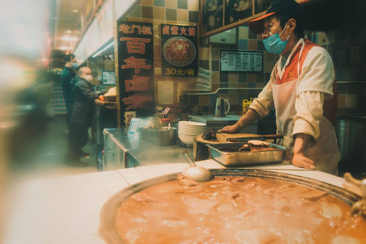 Isang street vendor sa likod ng isang nagsusugong kaldero ng hotpot sa isang food stall sa Chengdu, na may mga karatulang nakasulat sa Chinese at isang tiled na dingding.
