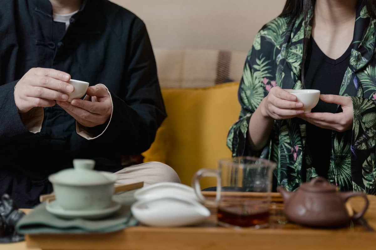 Zwei Personen, die an einem Tisch sitzen und kleine Teetassen halten, mit einem traditionellen Teeservice, Gaiwan und Teekessel vor ihnen.