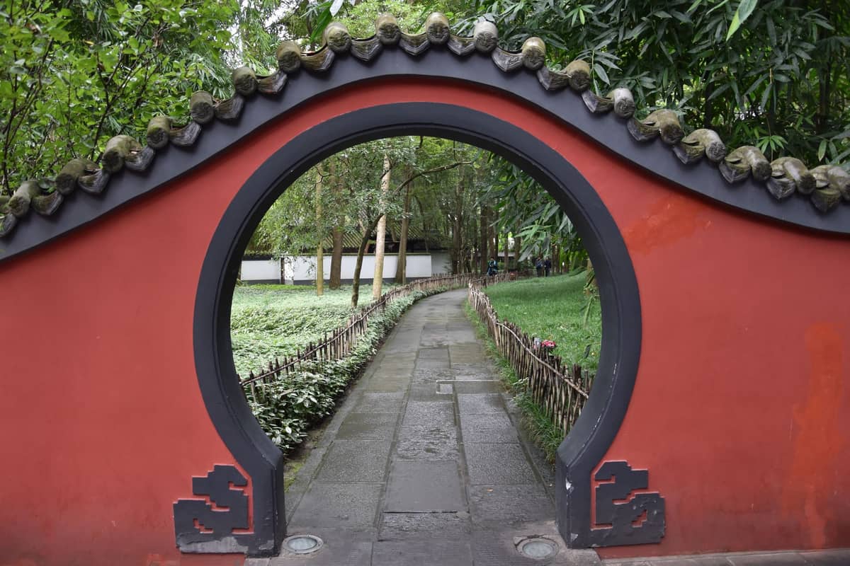 Rotes traditionelles chinesisches Mondtor, das zu einem Steinweg durch grüne Gärten und Bambus führt und auf eine historische Stätte in Sichuan hindeutet.