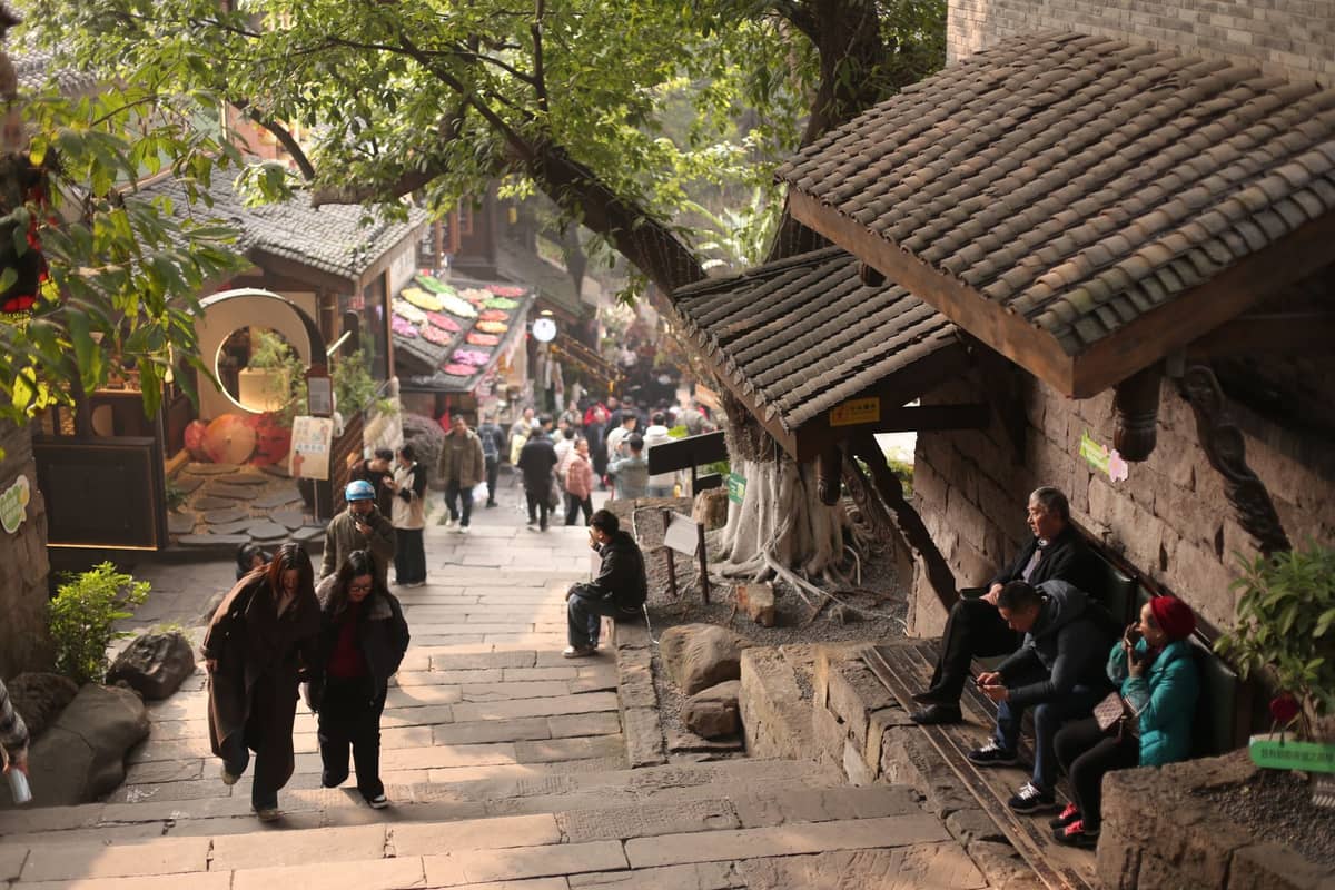 Eine lebhafte alte Steinstraße in einer historischen chinesischen Stadt nahe Chengdu, mit spazierenden Familien und Menschen, die sich neben traditionellen Gebäuden entspannen.