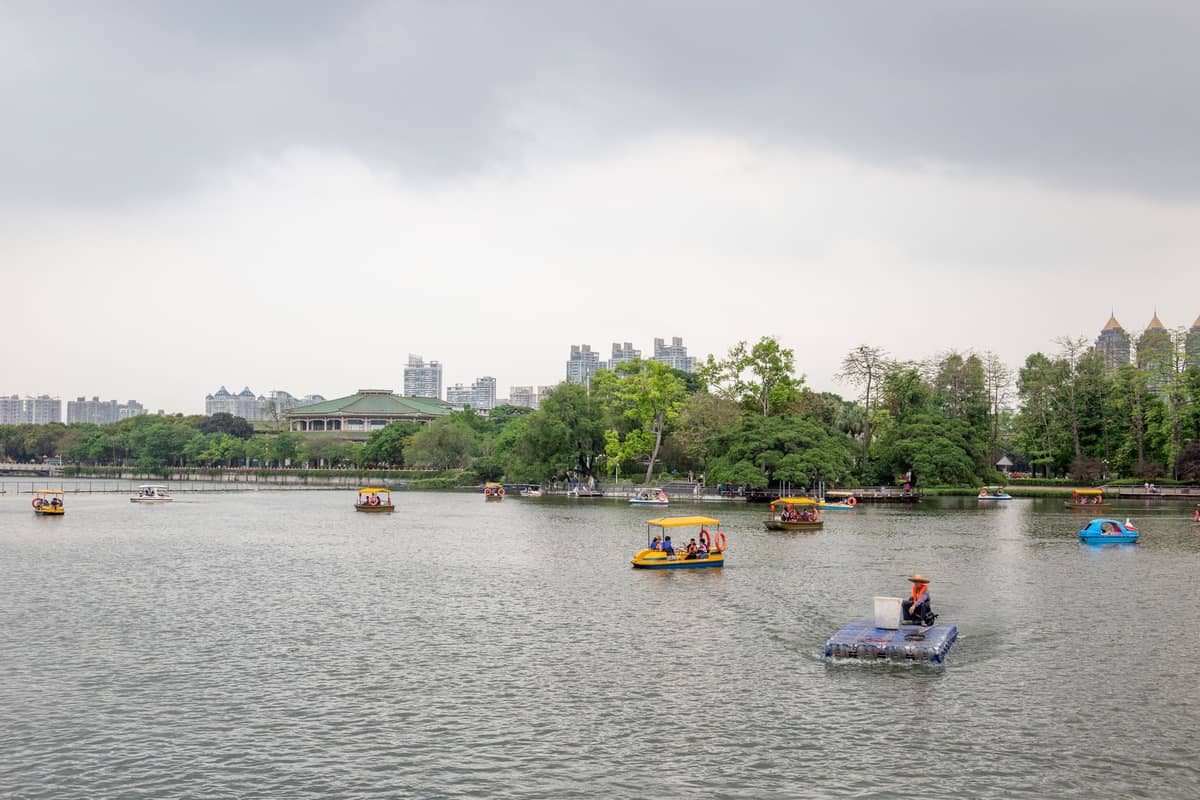 Menschen in bunten Tretbooten auf einem großen See in einem Stadtpark mit Bäumen und Hochhäusern im Hintergrund.