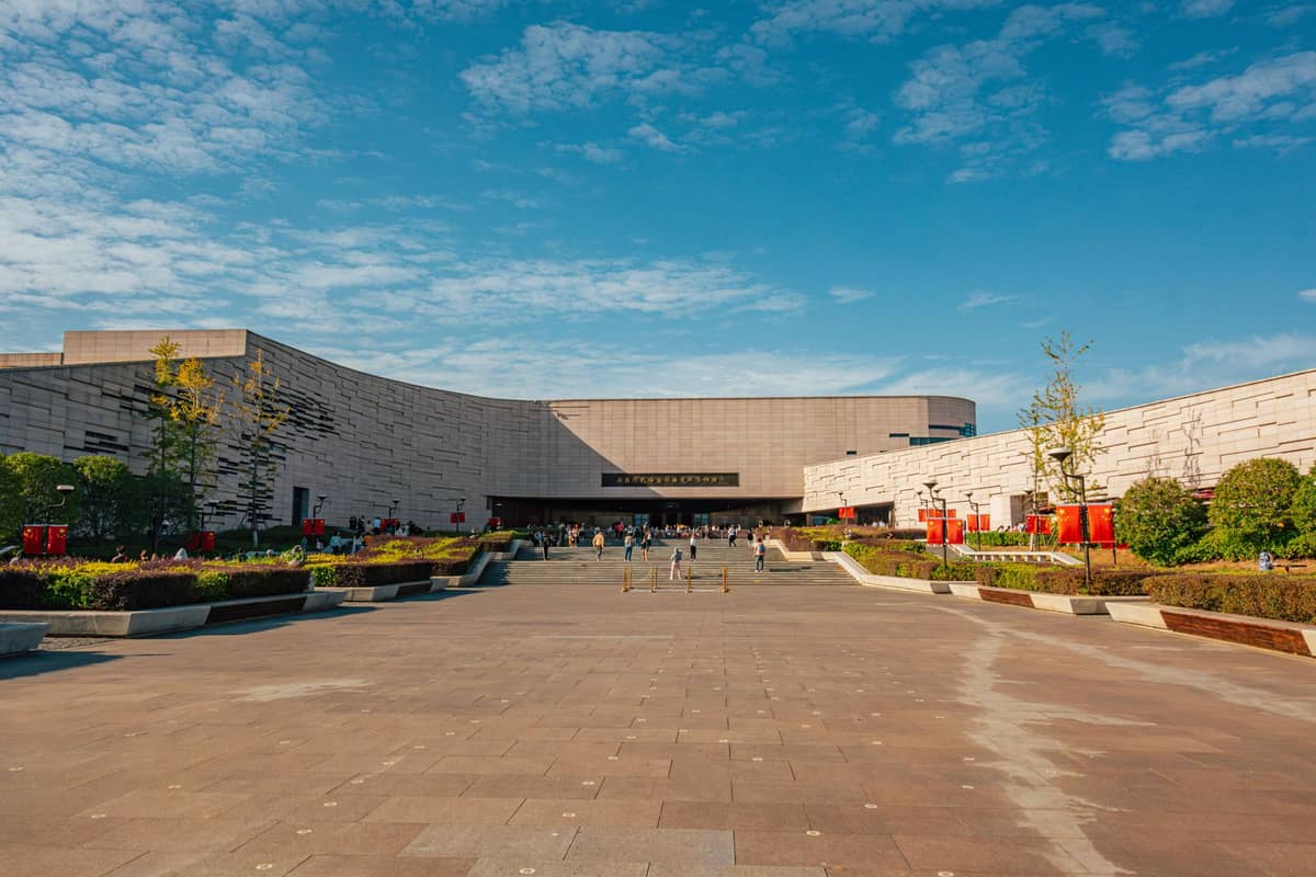 Weitwinkelansicht des modernen Eingangs des Sanxingdui-Museums in Guanghan mit Besuchern, blauem Himmel und gepflegten Gartenanlagen.