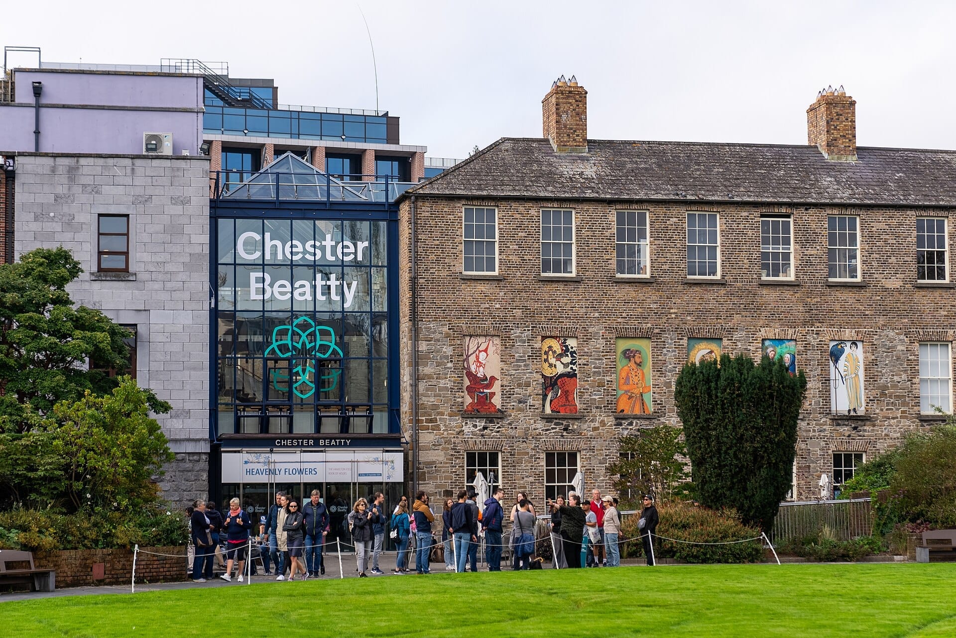 Vista esterna della Chester Beatty Library a Dublino con ingresso in vetro moderno, storici muri in mattoni, pannelli artistici colorati e visitatori in attesa all'esterno.