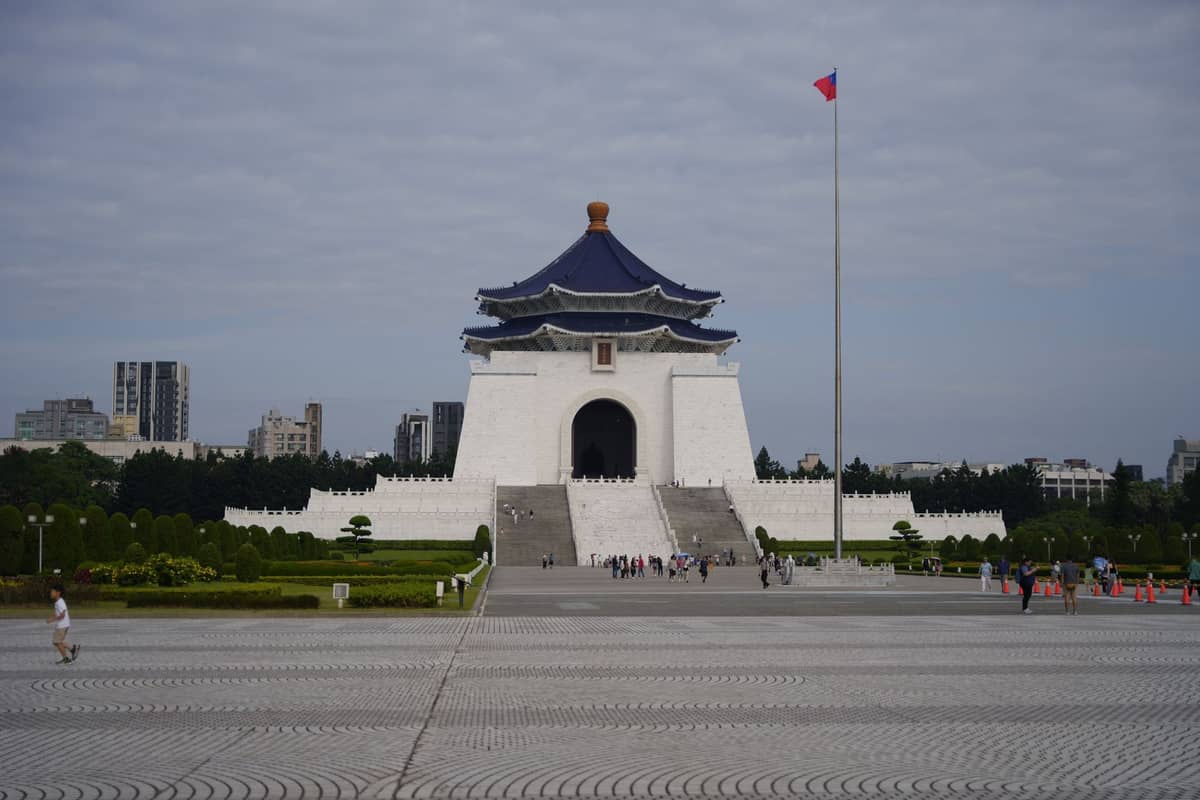 Frontansicht der Chiang-Kai-shek-Gedächtnishalle in Taipeh mit weißen Mauern, blauem achteckigem Dach, hohem Fahnenmast und dem belebten Vorplatz vor der Stadtkulisse.