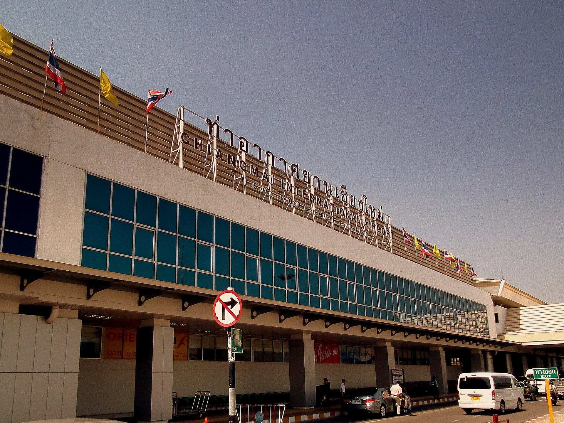 Vista exterior de la terminal del Aeropuerto Internacional de Chiang Mai con ventanas de vidrio, banderas y vehículos estacionados en la zona de recogida bajo un cielo despejado.